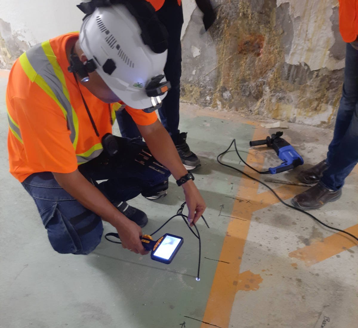 A construction worker in an orange safety shirt, white helmet, and knee pads is using a blue handheld device feeding a borescope inspection camera into the concrete floor below.