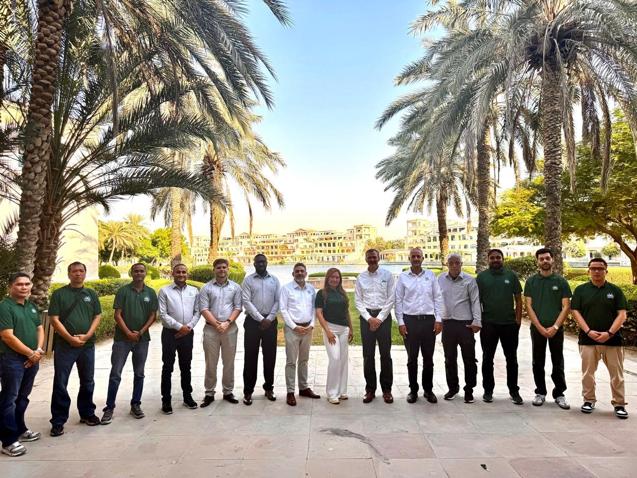 A group of 13 people standing outdoors in front of palm trees, with a large building and a fountain in the background, during daytime.