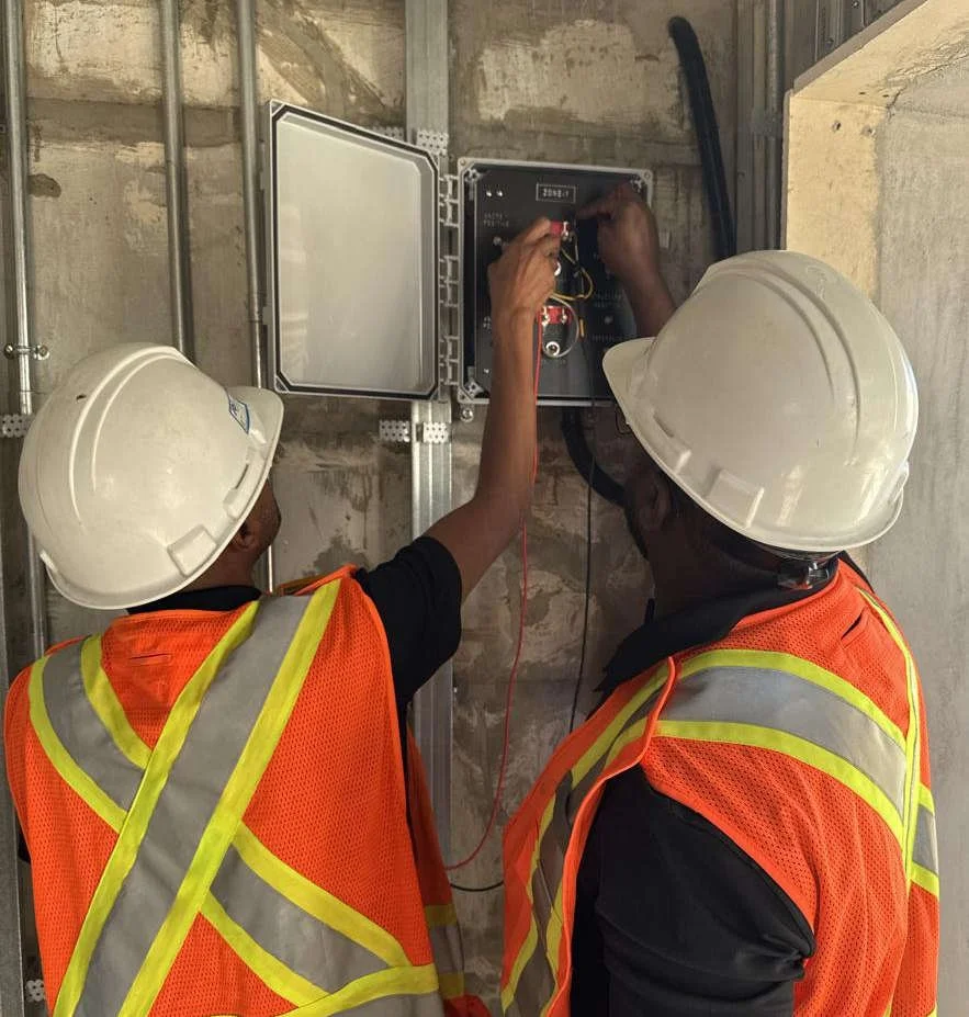 Two construction workers wearing safety helmets and orange safety vests working on an monitoring box at a construction site.