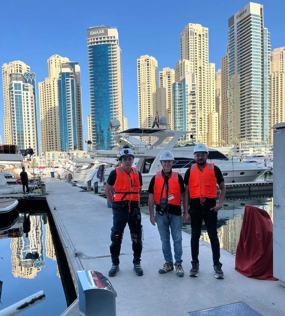 Three men wearing helmets and orange safety vests standing on a dock with boats, and tall city buildings in the background.