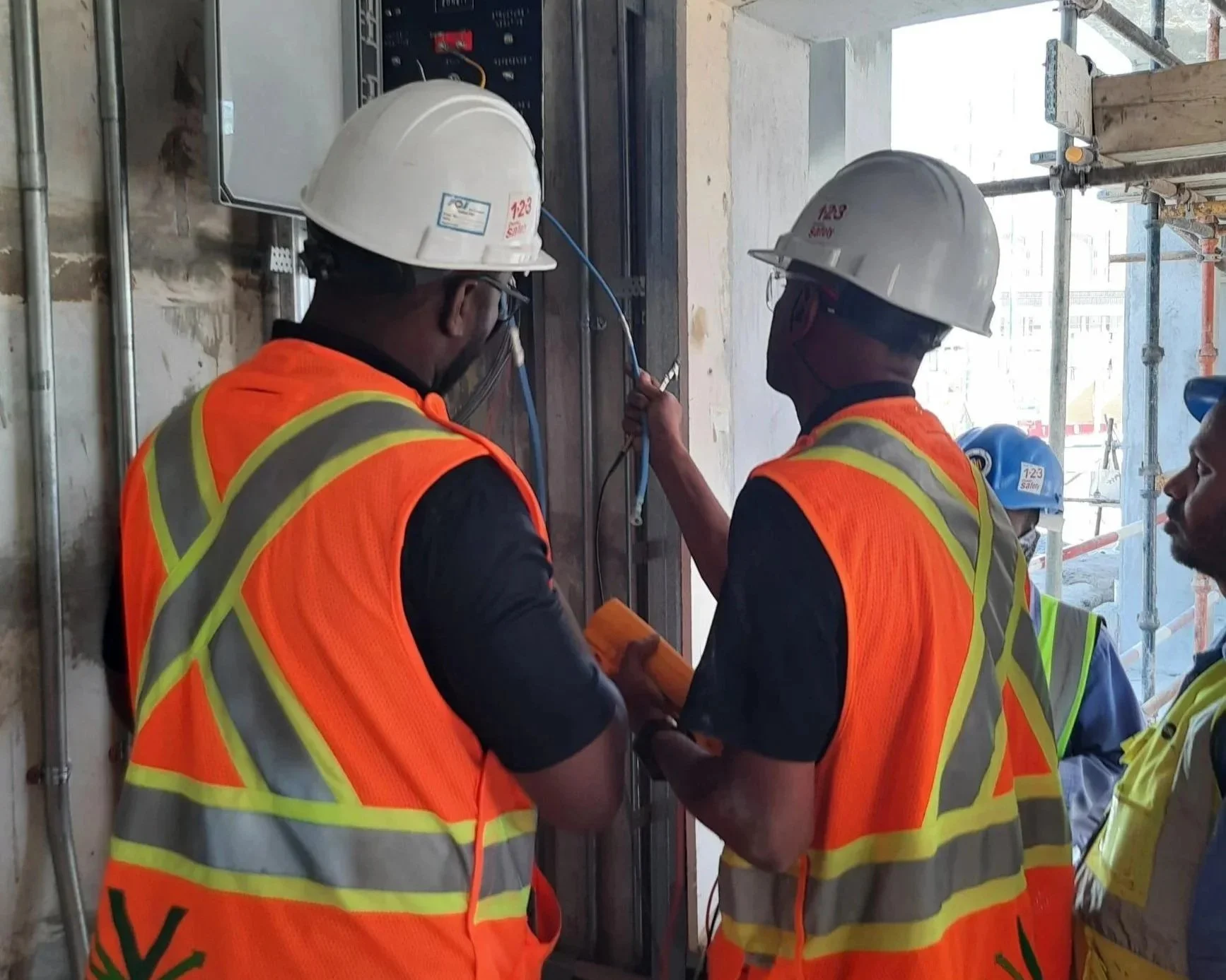 Construction workers wearing safety helmets and orange reflective vests working on monitoring box inside a building under construction.