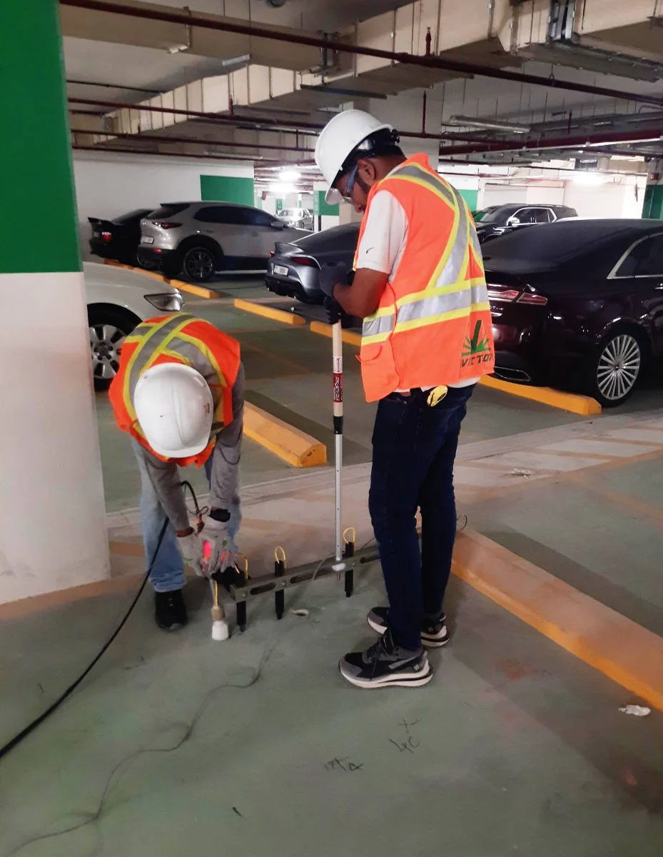 Two construction workers in safety vests and helmets working on underground car park, installing equipment on concrete floor near parked cars.