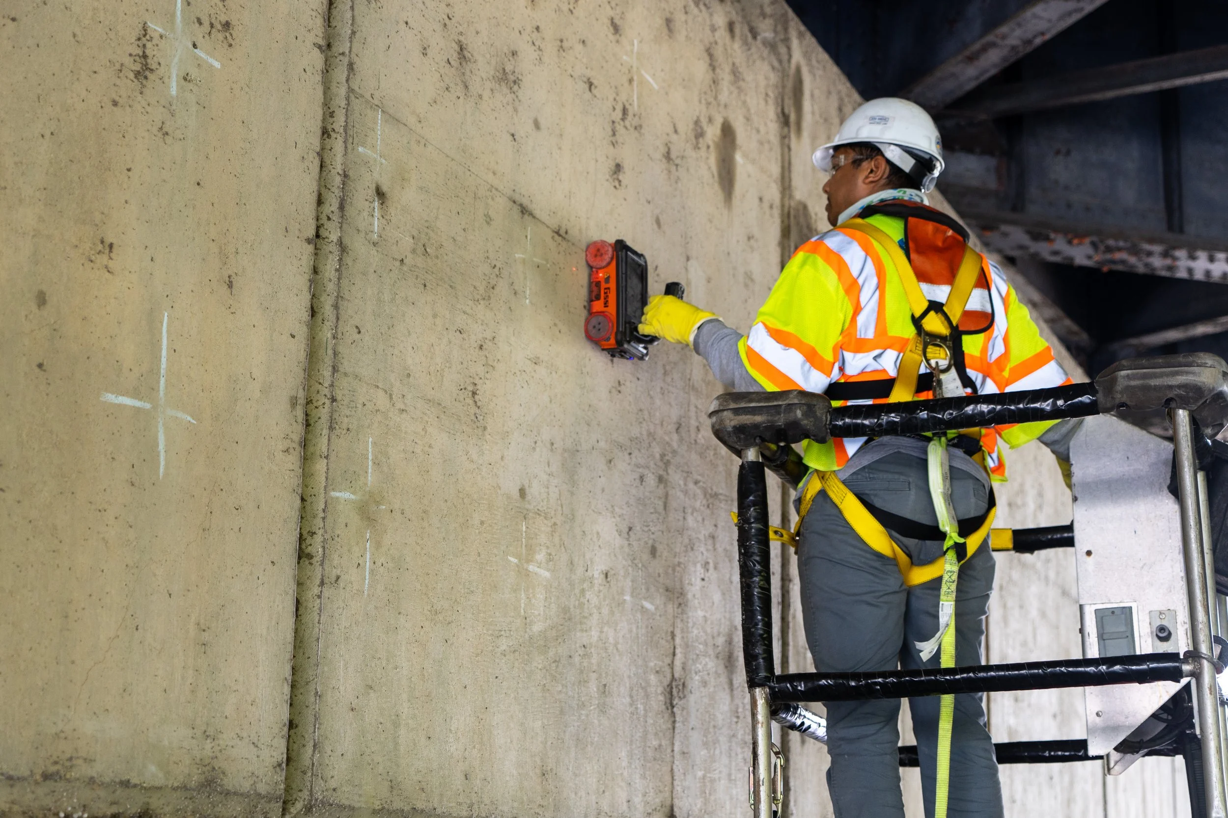 A construction worker wearing a white safety helmet, yellow safety gloves, and a high-visibility jacket kneels on a scaffold while using a handheld GPR device to map out reinforcement on a concrete wall.