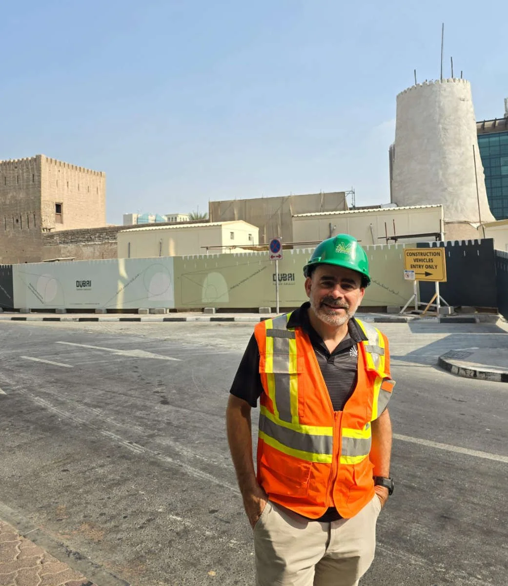 Construction worker standing on a street in front of a construction site with Dubai branding. The worker is wearing a green safety helmet, an orange reflective safety vest, and casual pants, smiling at the camera.