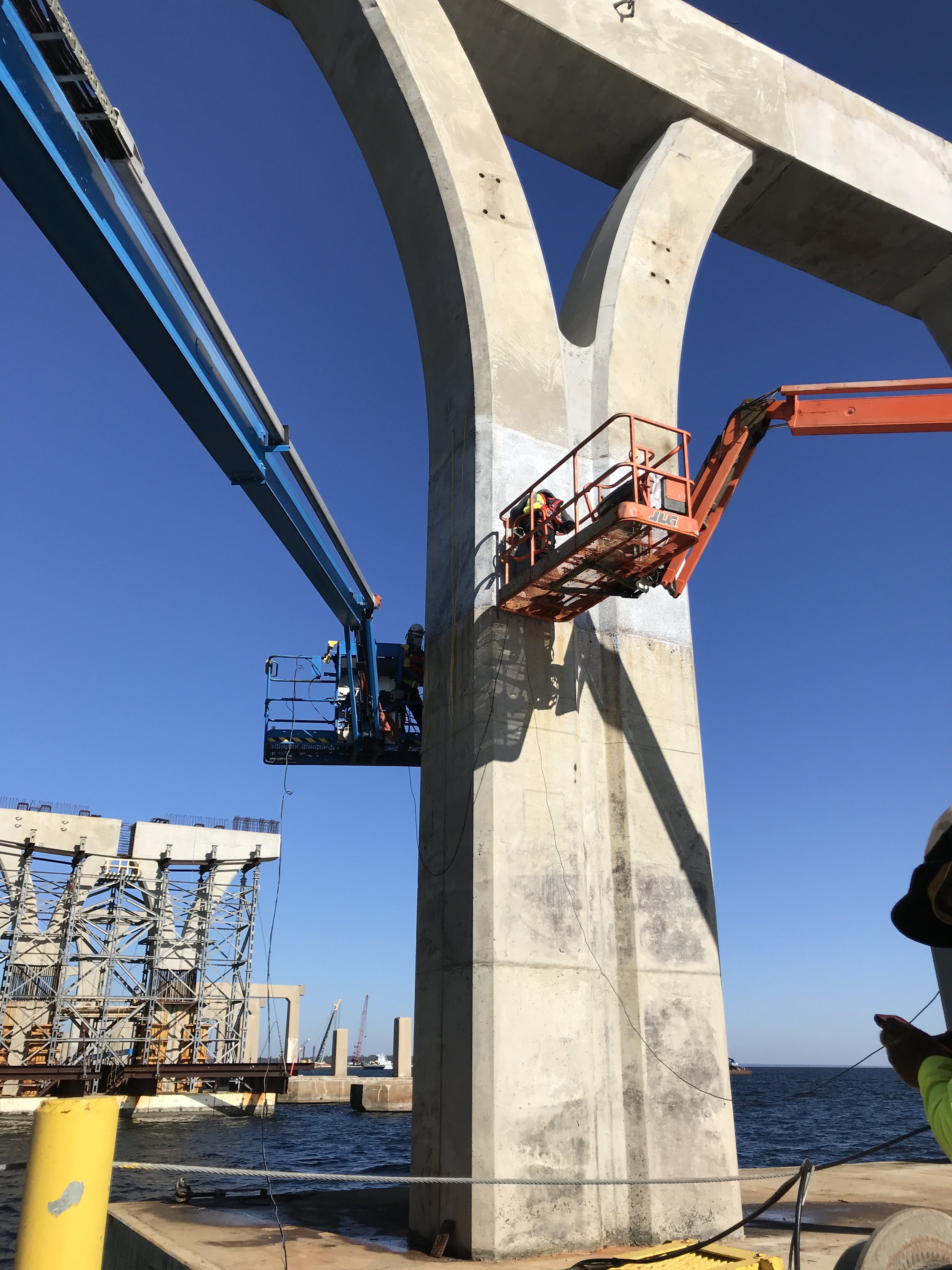 Construction workers on orange lifts working on a large concrete bridge support near the water, with a clear blue sky and a crane in the background.