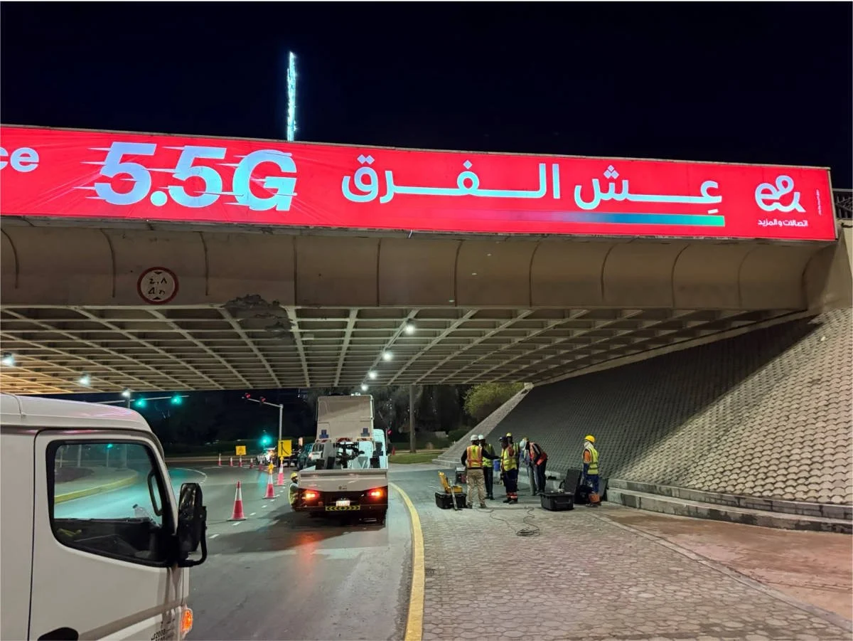 Under a bridge, several construction workers in safety vests and helmets are working near equipment and tools at night. There are parked vehicles, cones, and traffic lights visible.