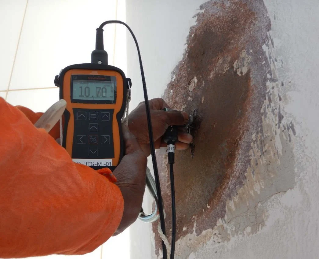 A person testing the moisture level of a wall with a digital moisture meter. The device displays a reading of 10.78% and is connected to a probe inserted into a damaged area of the wall.
