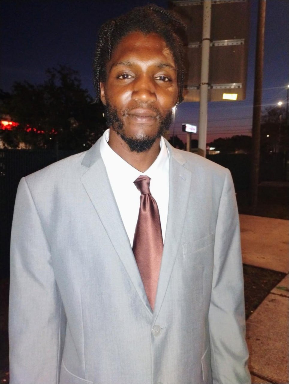 A man wearing a gray suit, white shirt, and brown tie standing outdoors during twilight.