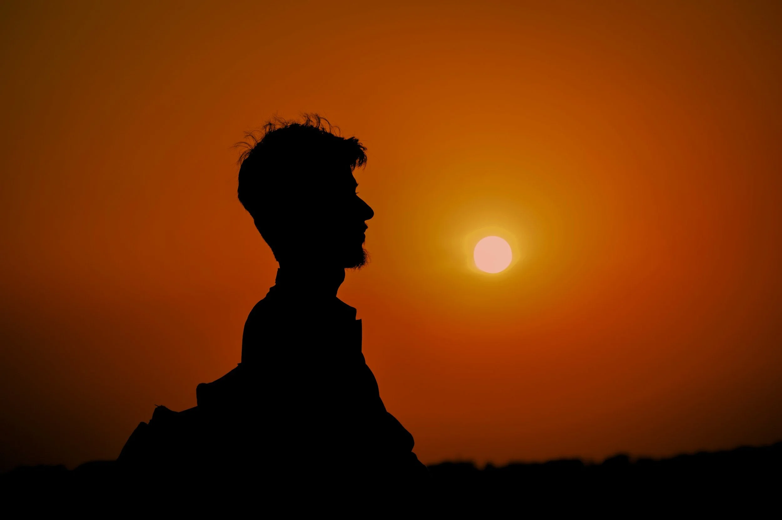 Silhouette of a person with a beard and messy hair standing beside a mountain, with the sun setting behind them, creating an orange sky.