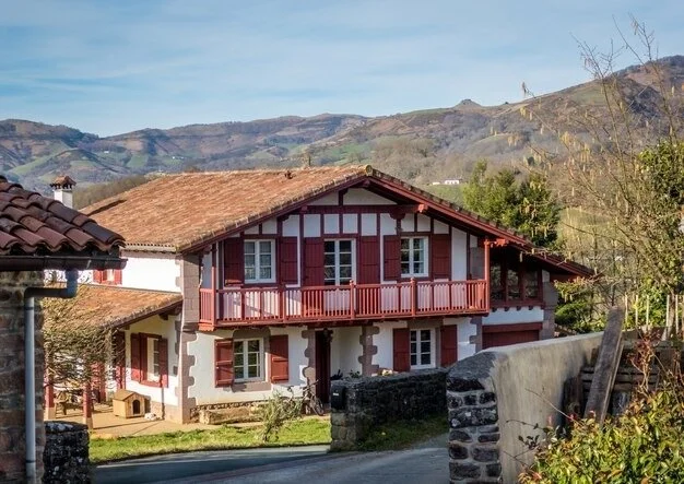 A traditional house with a red-tiled roof and red window shutters, located in a rural area with hills in the background.