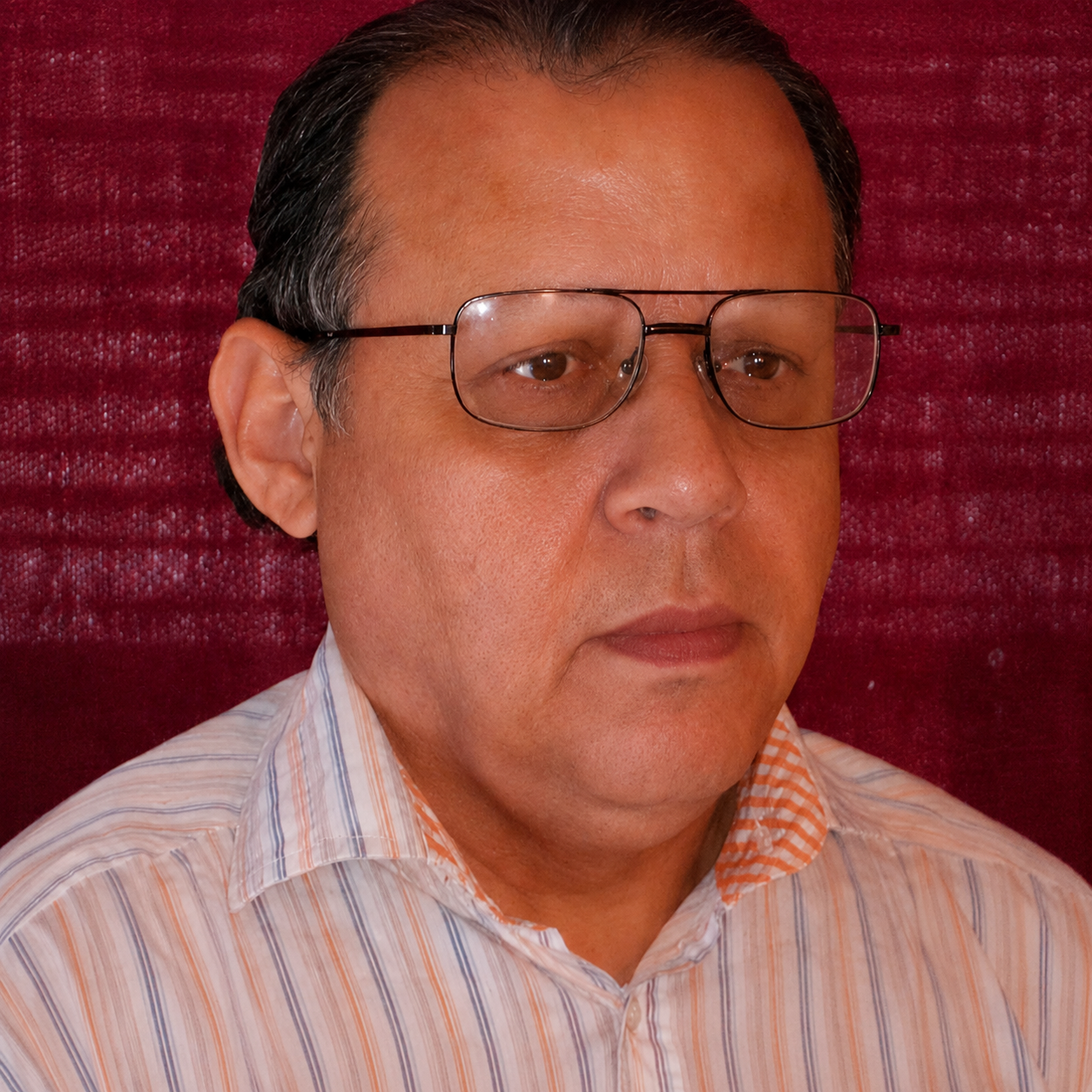 A middle-aged man with glasses wearing a striped collared shirt in front of a red textured background.