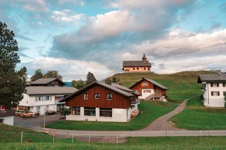 A scenic view of a small village with several houses on a grassy hillside, featuring a church or chapel on top of the hill, under a partly cloudy sky.
