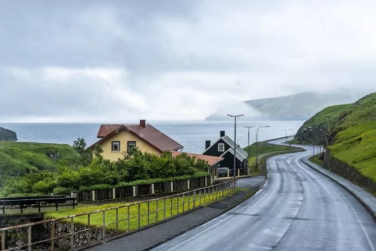 A winding coastal road with green hills, houses, and a view of the ocean under a cloudy sky.