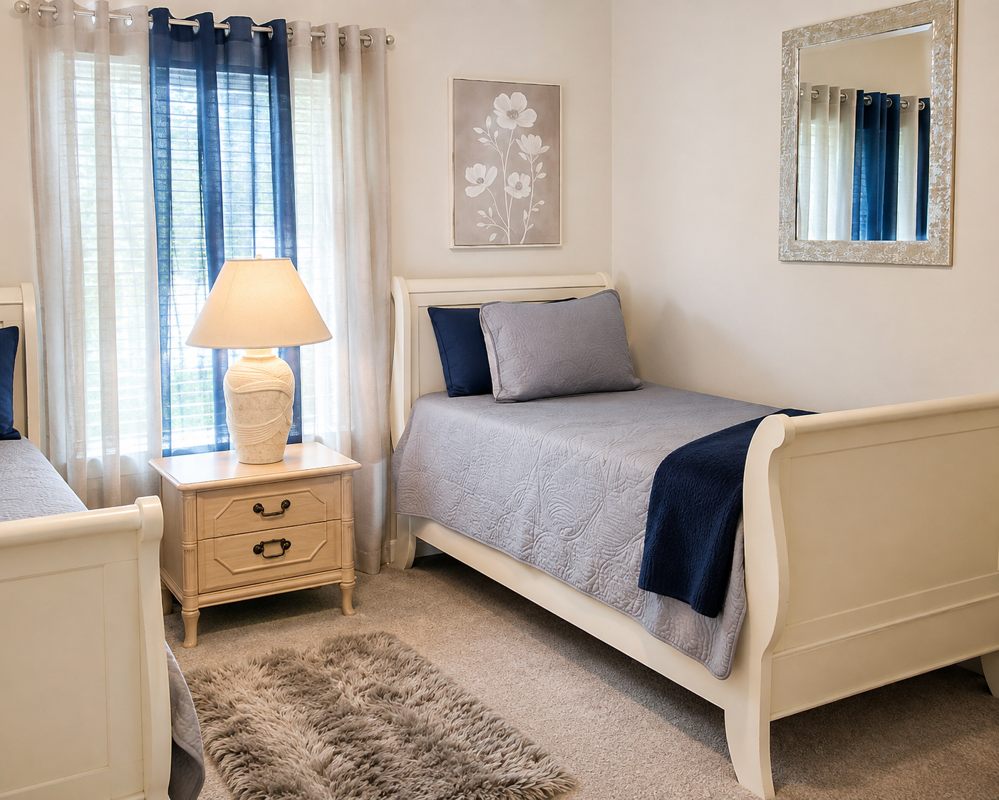 A cozy bedroom with two white beds with gray and navy blue bedding, a nightstand with a beige lamp, a window with navy and beige curtains, a floral wall art, a mirror, and a plush gray rug.