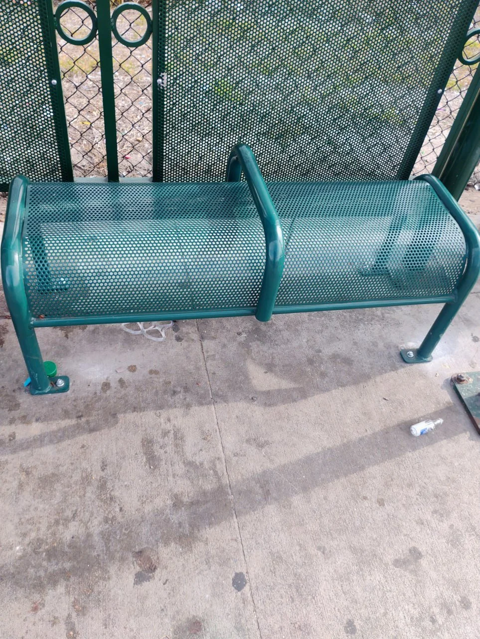 Empty green metal park bench on concrete ground with a metal fence backdrop and scattered trash.
