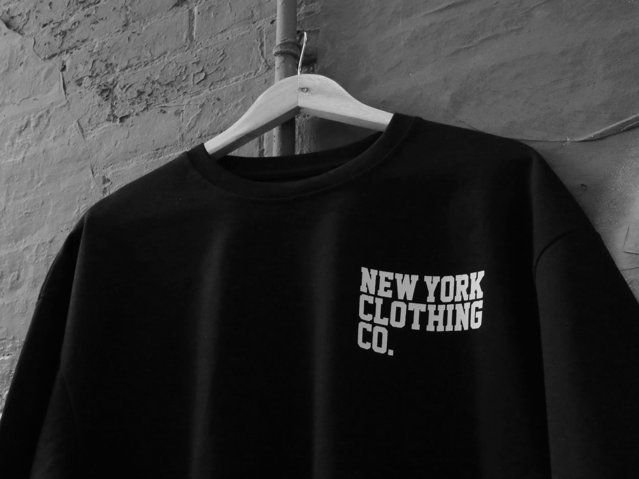 A black t-shirt hanging on a wooden hanger on a pipe against a brick wall.