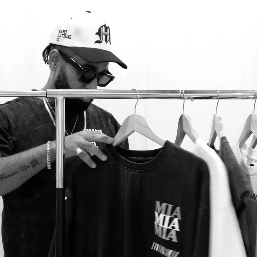 Christian Noa scanning through a rack wearing sunglasses and a cap, looking at black t-shirts on a clothing rack.