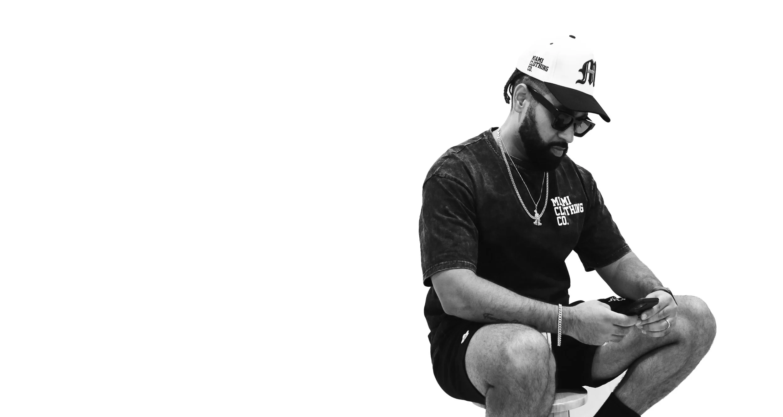 A man with a beard wearing sunglasses, a baseball cap, a T-shirt with 'Mimi Clothing Co.' written on it, and shorts, is sitting on a stool looking at his phone.