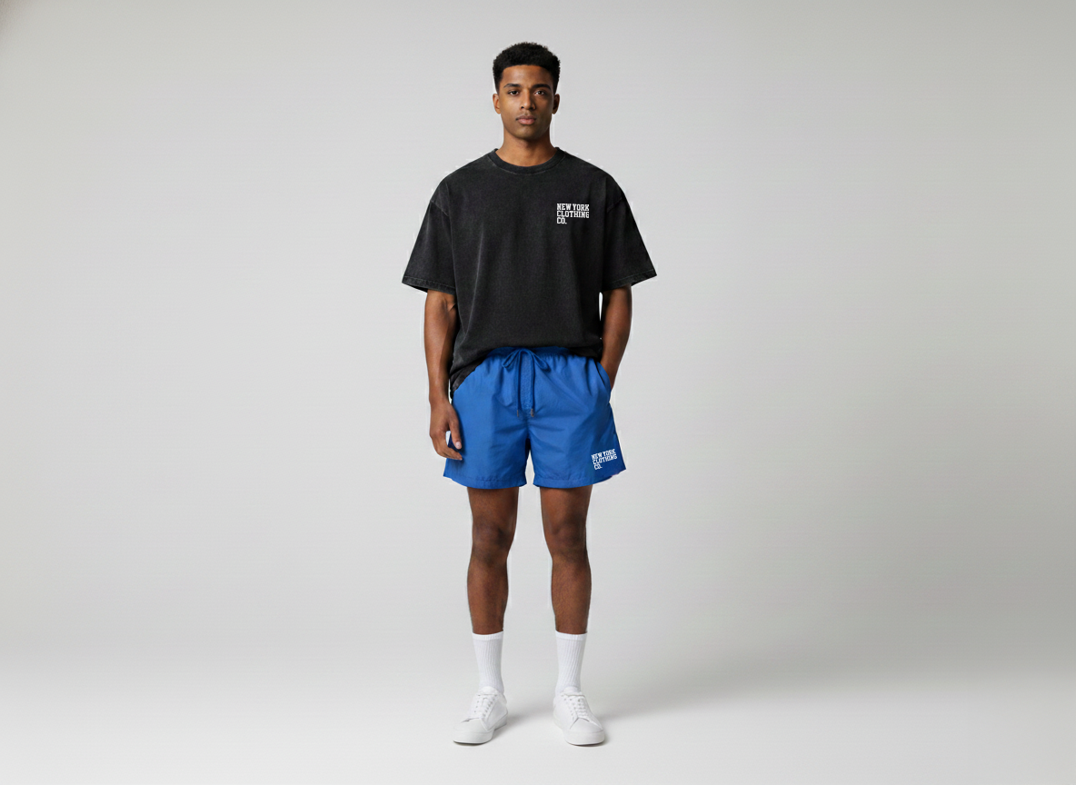 Young man in black T-shirt, blue shorts, white socks, and white sneakers, standing with hands in pockets against a plain light-colored background.
