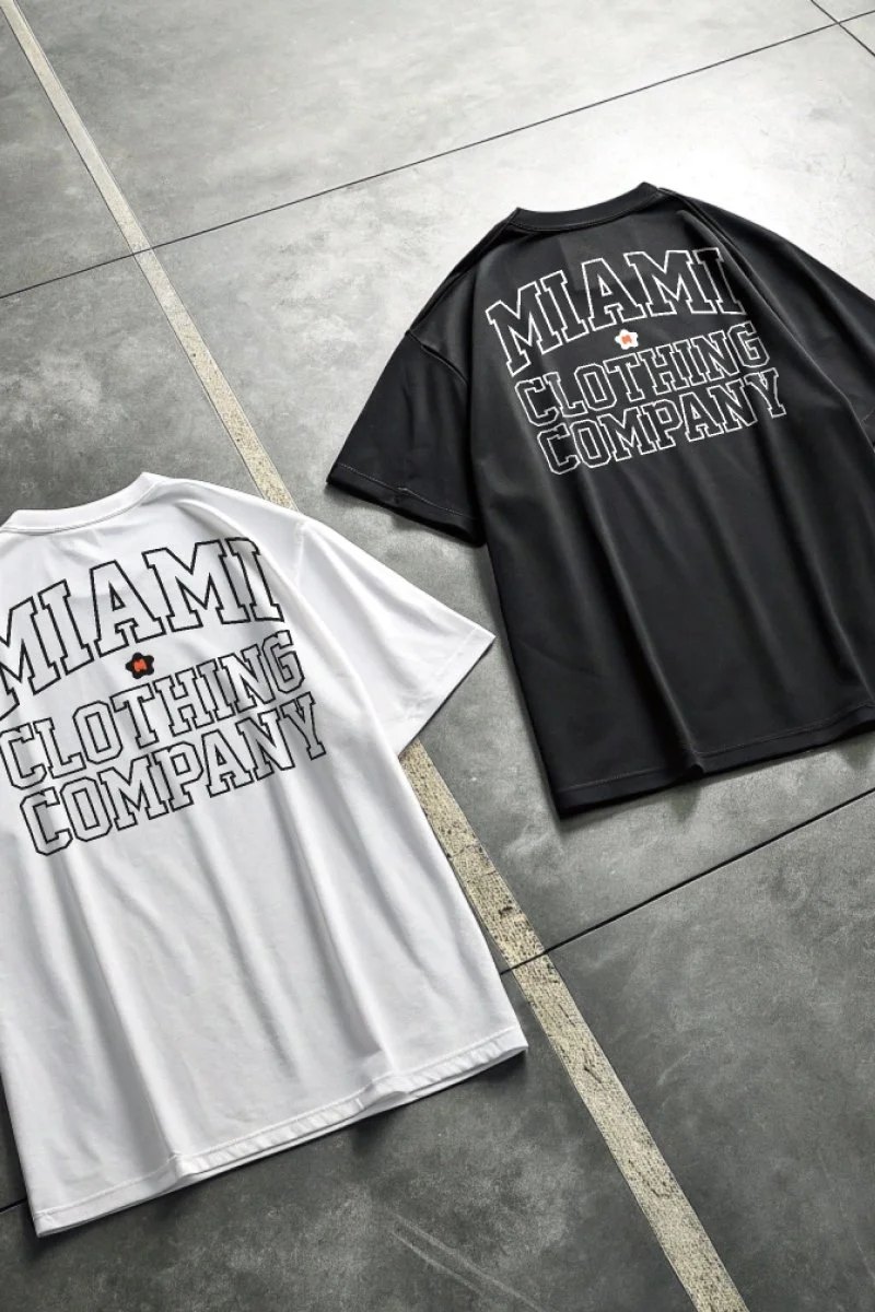 Two T-shirts, one white and one black, laid flat on a gray tiled floor. Both shirts have the words "Miami Clothing Company" printed on the back with a small orange flower above the text.