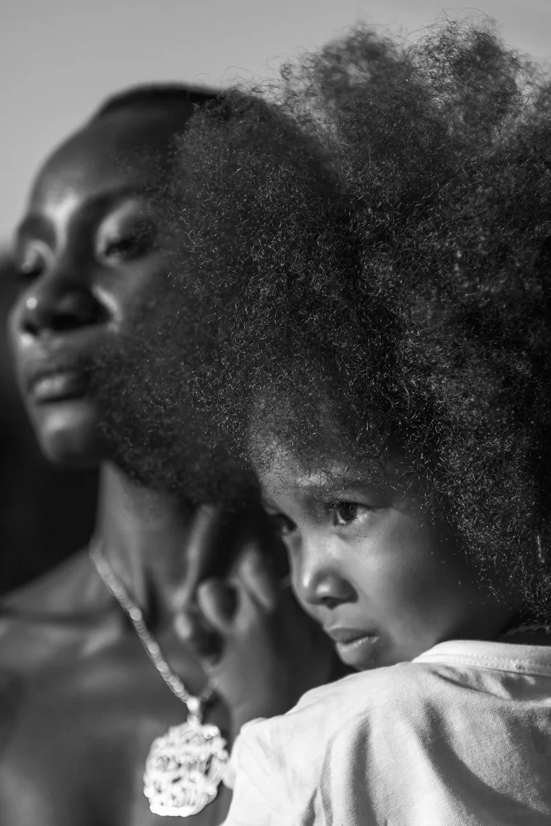 A woman with natural curly hair looking to the side and a young girl with similar curly hair resting her head on the woman's shoulder, both facing slightly to the left.