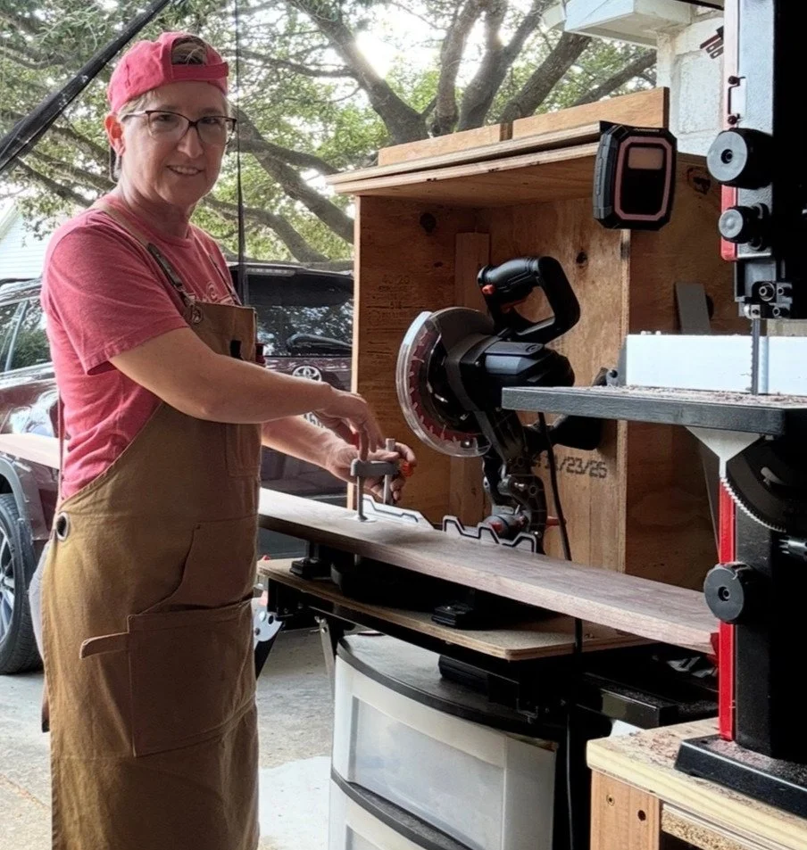 A woman in a red cap and glasses using a miter saw in a woodworking workshop, with a wooden fence and trees visible outside the window.