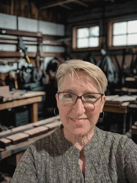 A woman with short blonde hair and glasses smiling in a woodworking shop with wooden workbenches and tools in the background.