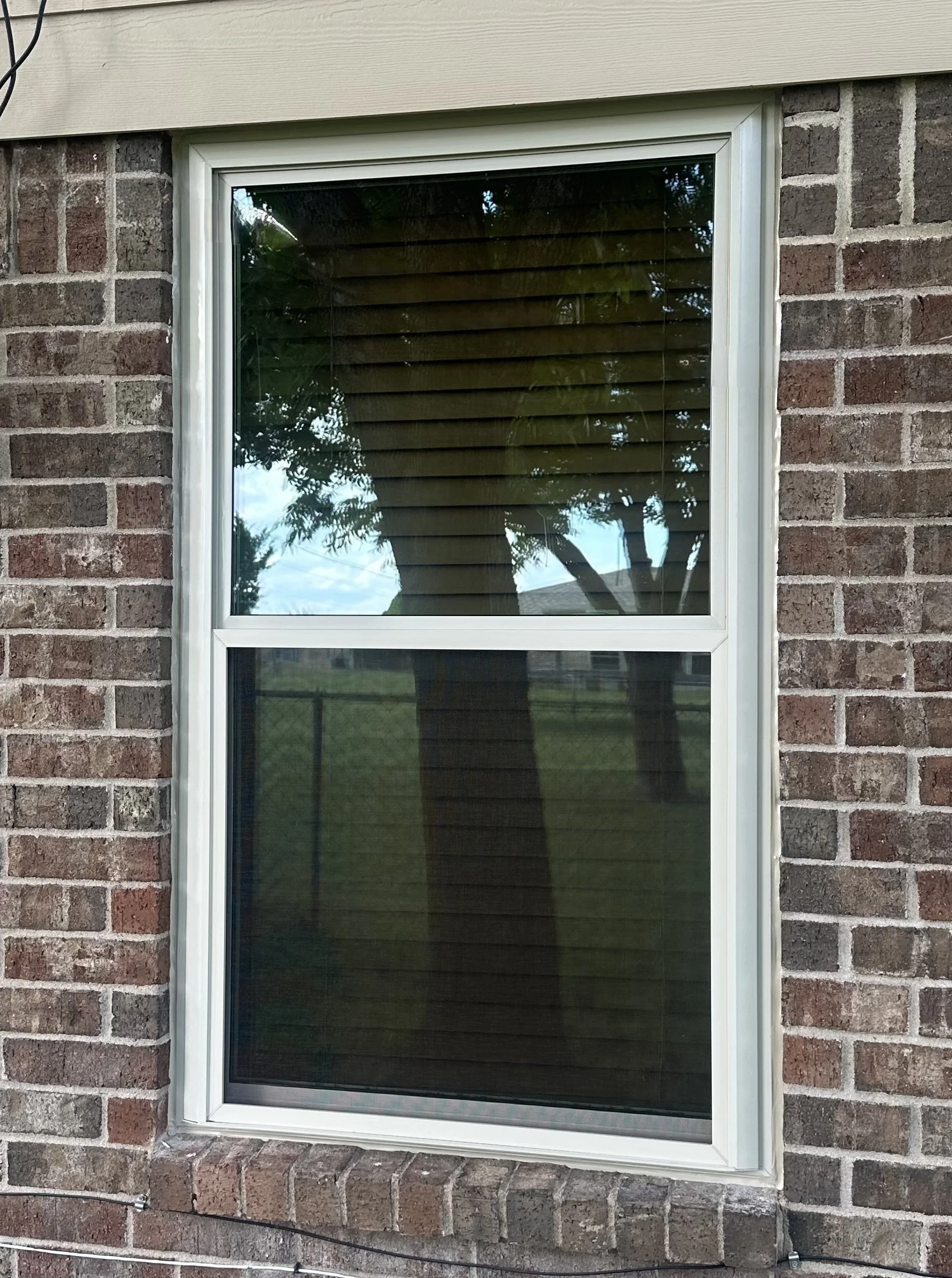A brick house wall with a double-pane window and white trim. Reflection of trees and sky can be seen on the window.