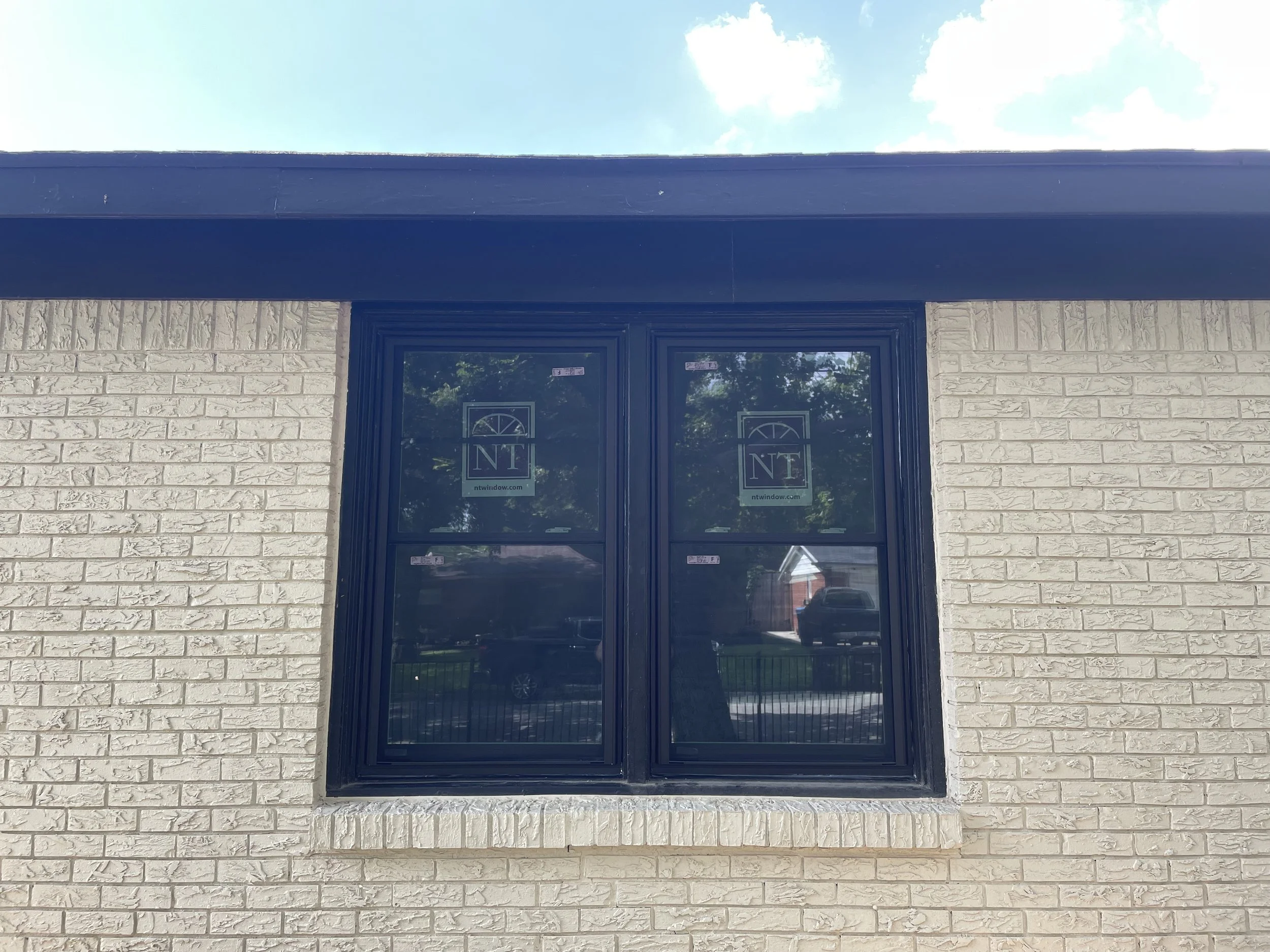 A storefront with large windows in a black frame, white brick walls on either side, and a blue awning above. The reflection of cars and trees can be seen in the windows, and the sky with clouds is visible above the building.