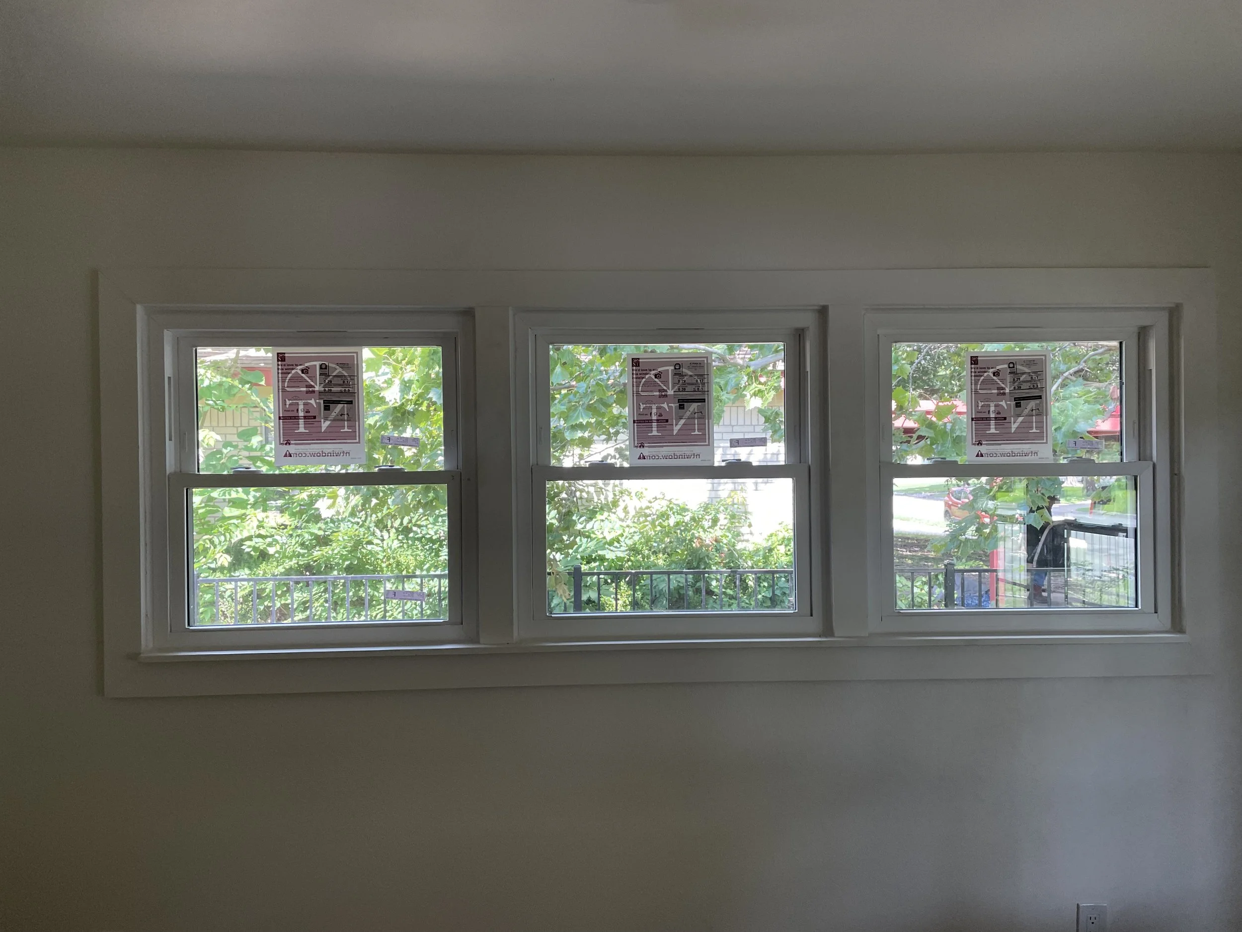 Interior view of a room with three large windows, each with a sticker indicating they are new and not to be opened yet, and outside visible trees and a street.
