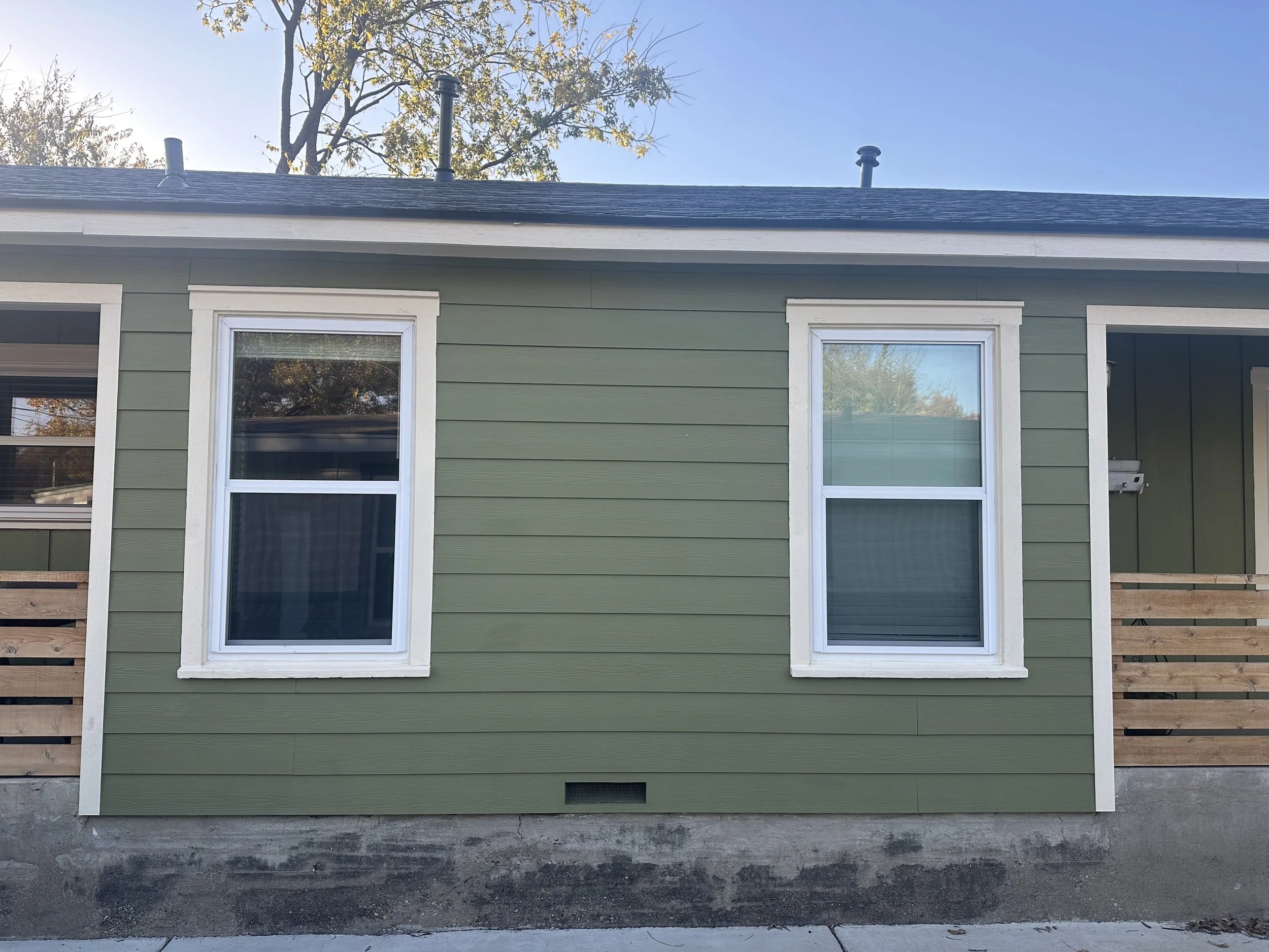 Exterior view of a house with green siding, two white-framed windows, and a black roof. There are two vents on the roof and a tall tree behind it. The house is partially under construction with a wooden fence on the side and a concrete foundation.