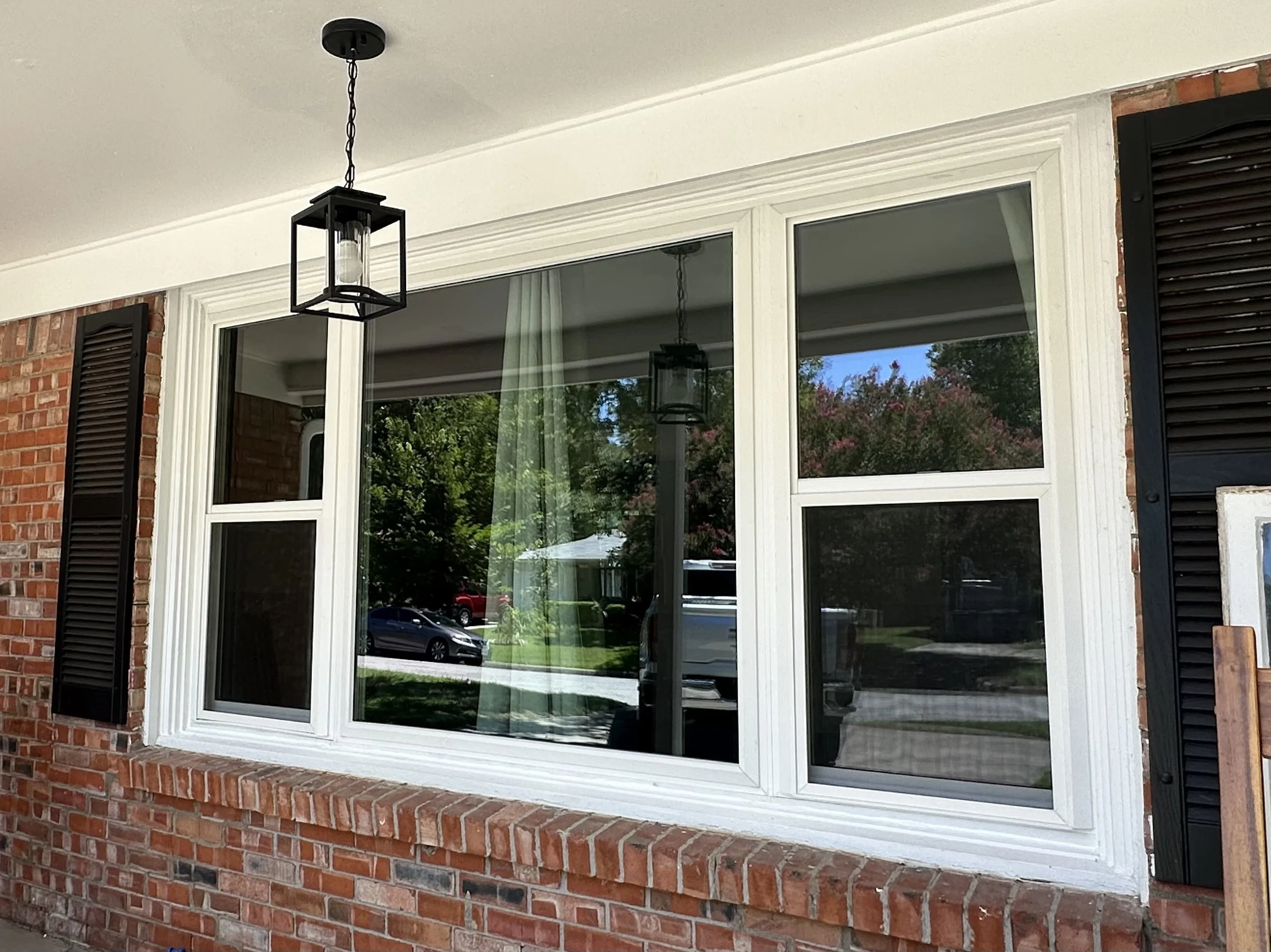 Exterior view of a house window with black shutters, a brick wall, and hanging black lantern-style outdoor lights, reflecting trees and a rotating umbrella outside.