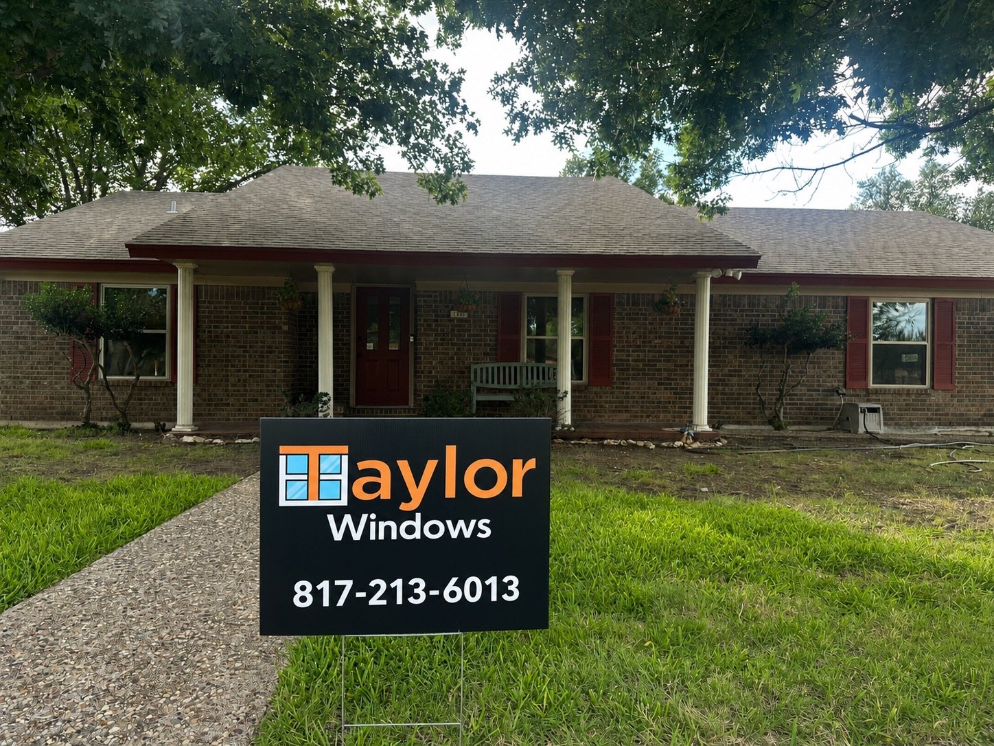Front view of a brick house with a porch, two trees, and a sign for Taylor Windows on a lawn.