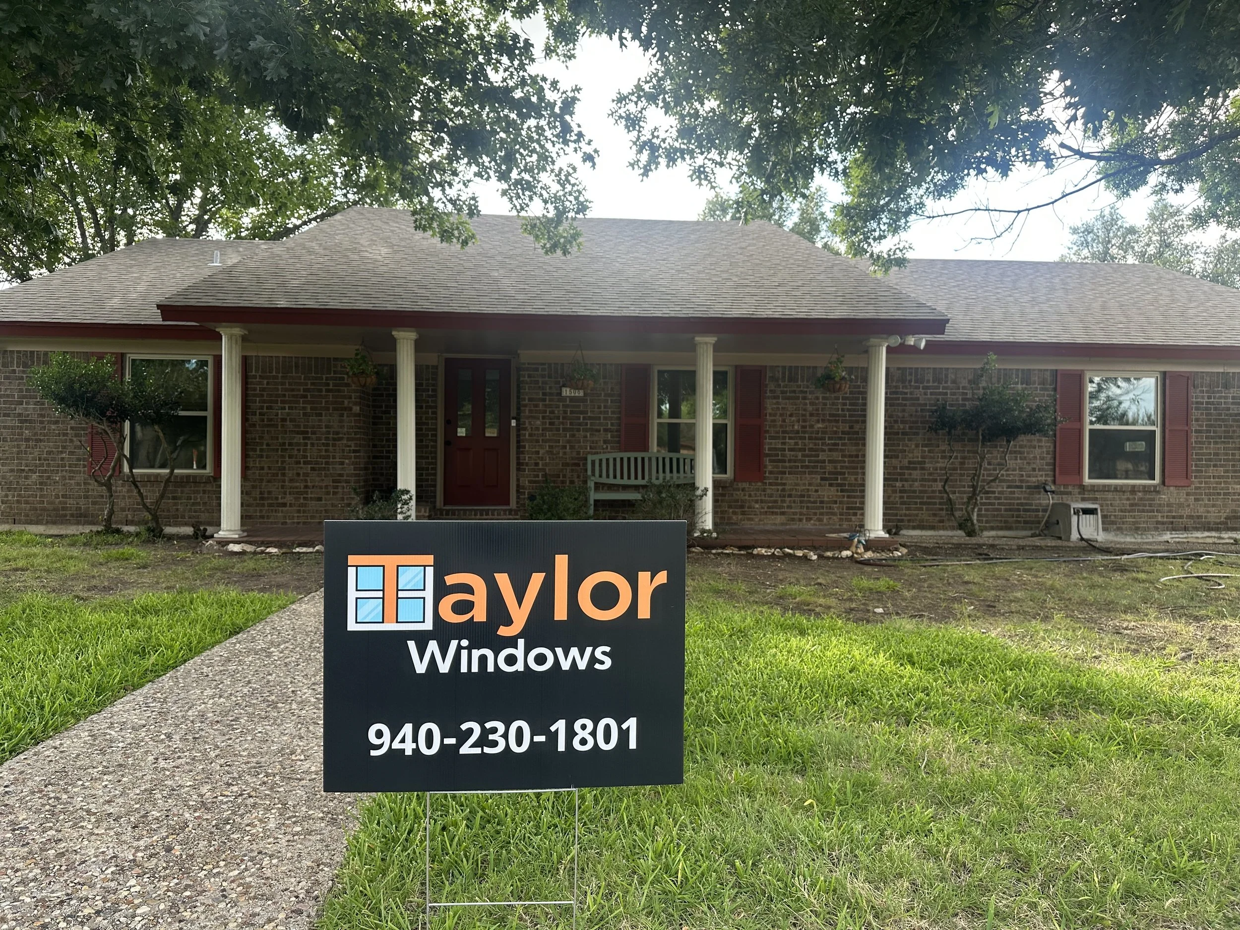 Front view of a brick house with a porch, two trees, and a sign for Taylor Windows on a lawn.
