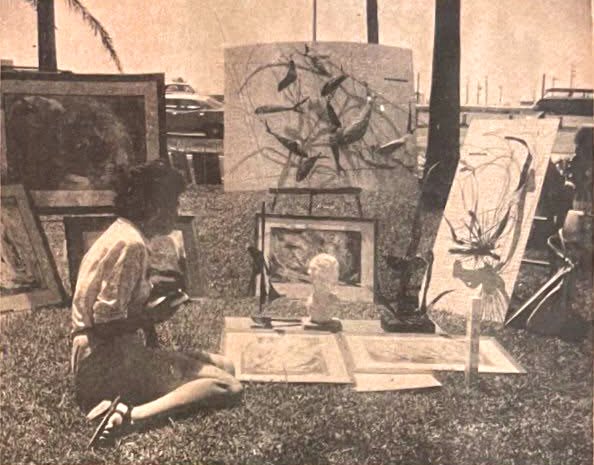 Woman sitting on grass at outdoor art display with paintings and artwork on display.