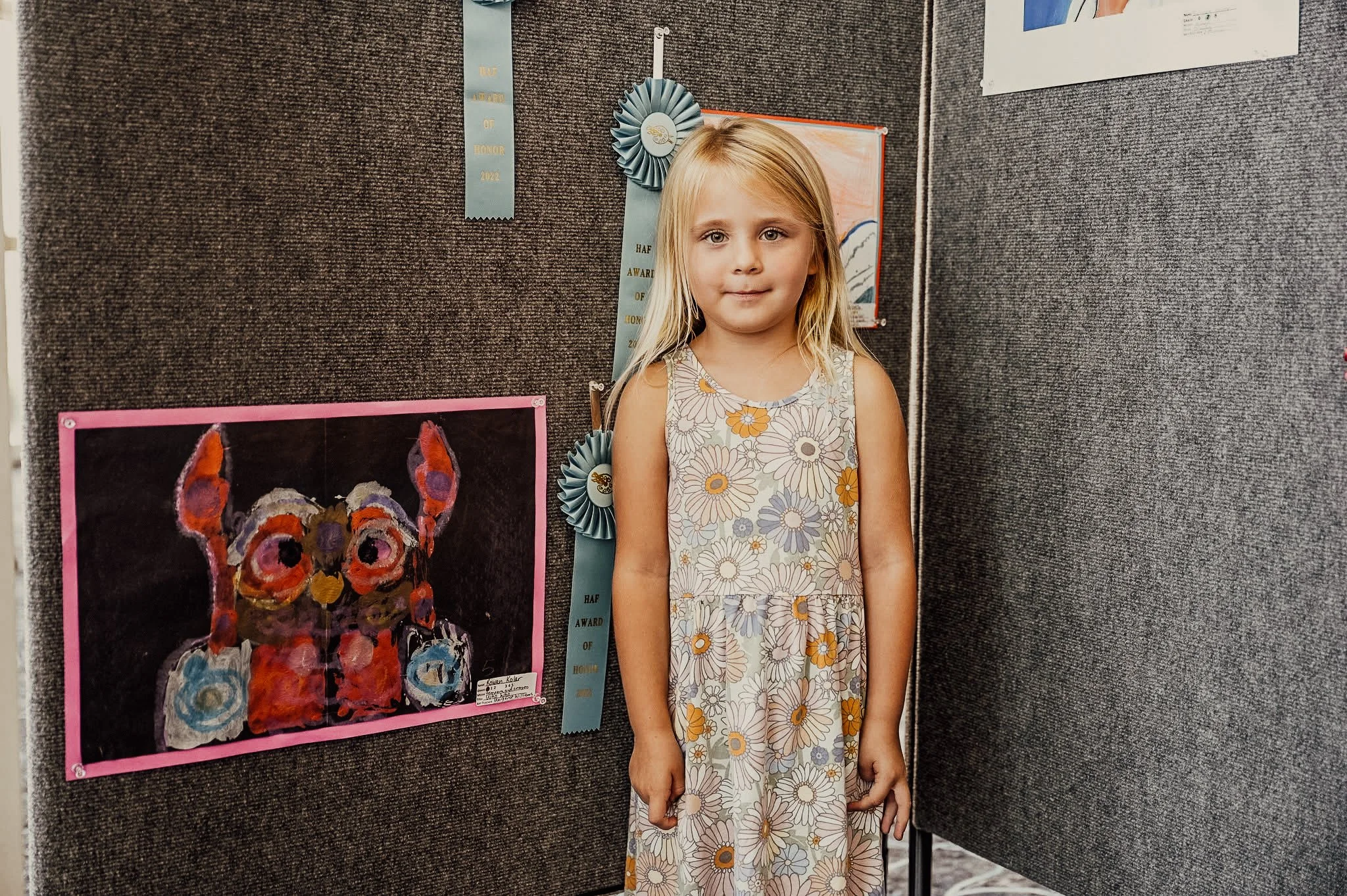A young girl with blonde hair wearing a sleeveless dress with a daisy pattern, standing in front of an art display with a colorful butterfly painting and award ribbons on a dark fabric background.