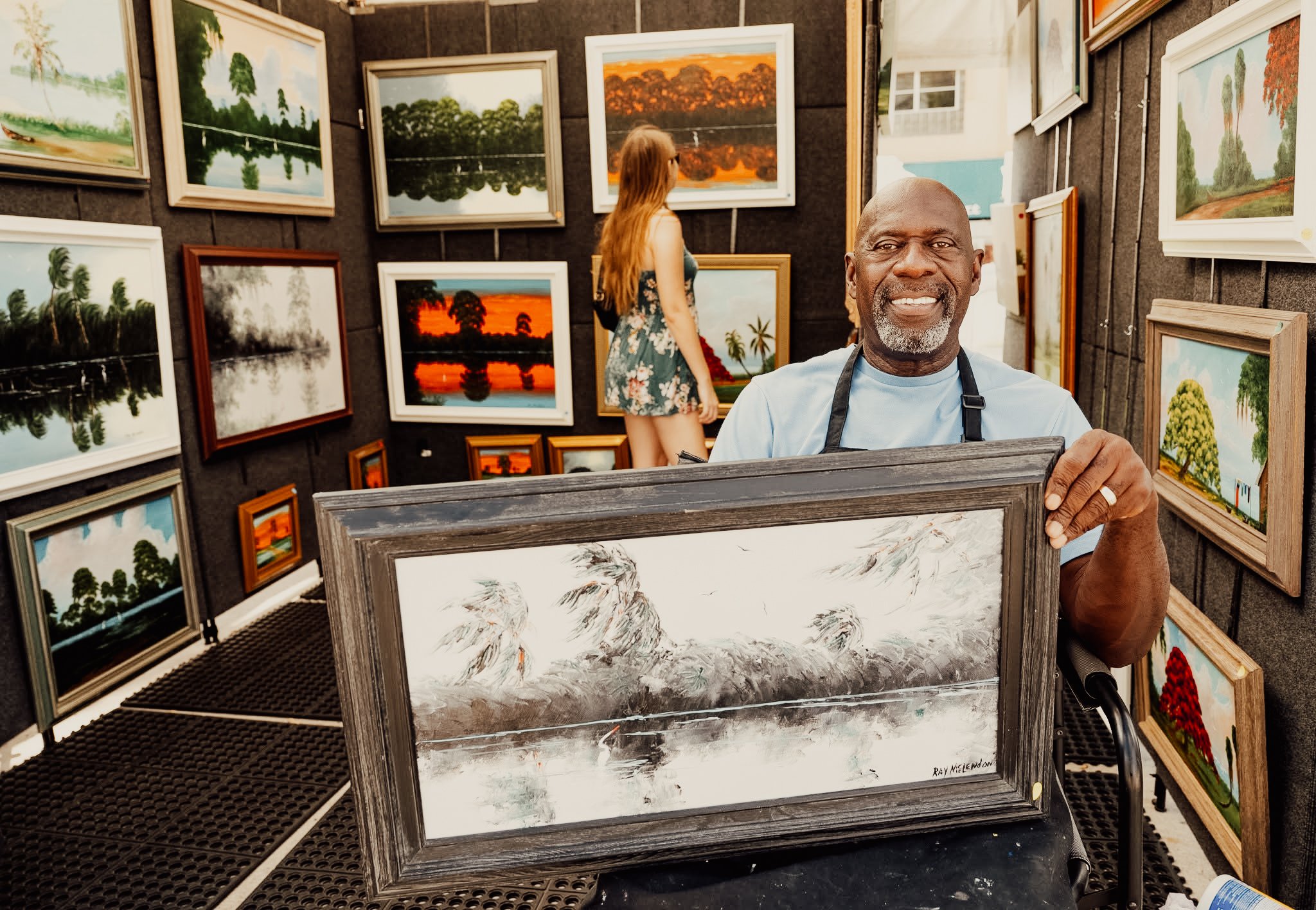 A man with a gray beard, smiling, holding a framed painting of a landscape, in an art vendor booth with various landscape paintings on the walls, and a woman with long reddish-brown hair looking at paintings in the background.
