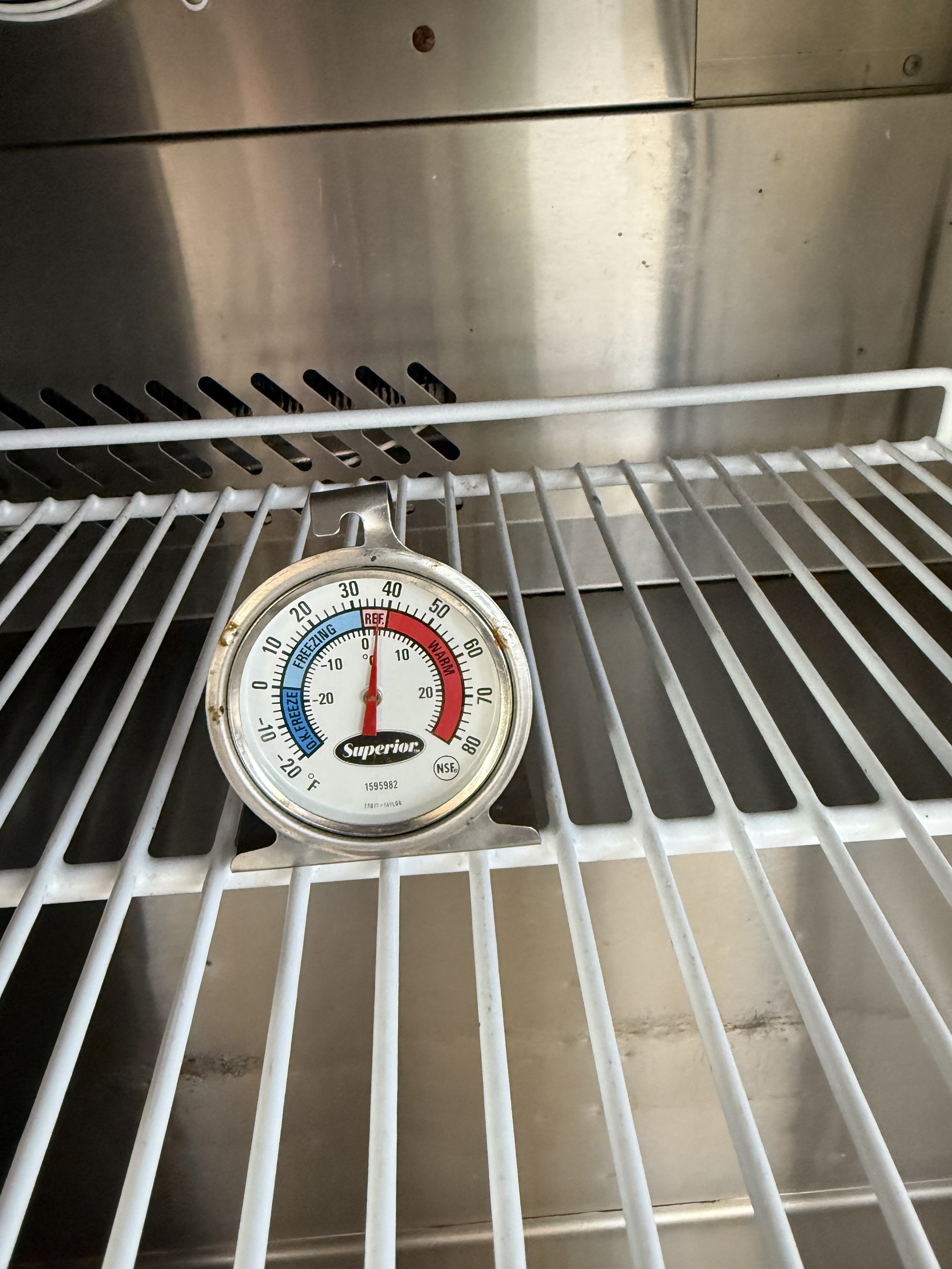 Inside of a refrigerator showing a temperature gauge on a wire rack, with a stainless steel back wall.