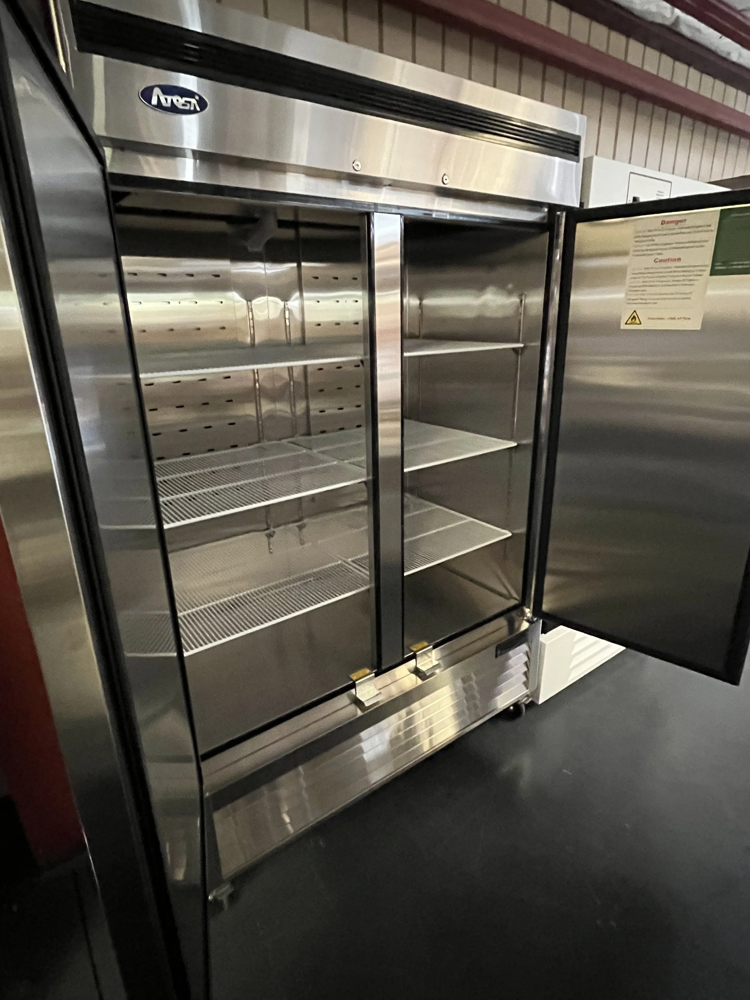 Empty stainless steel commercial refrigerator with glass door, shelves, and a caution sign, located in a storage or kitchen area.