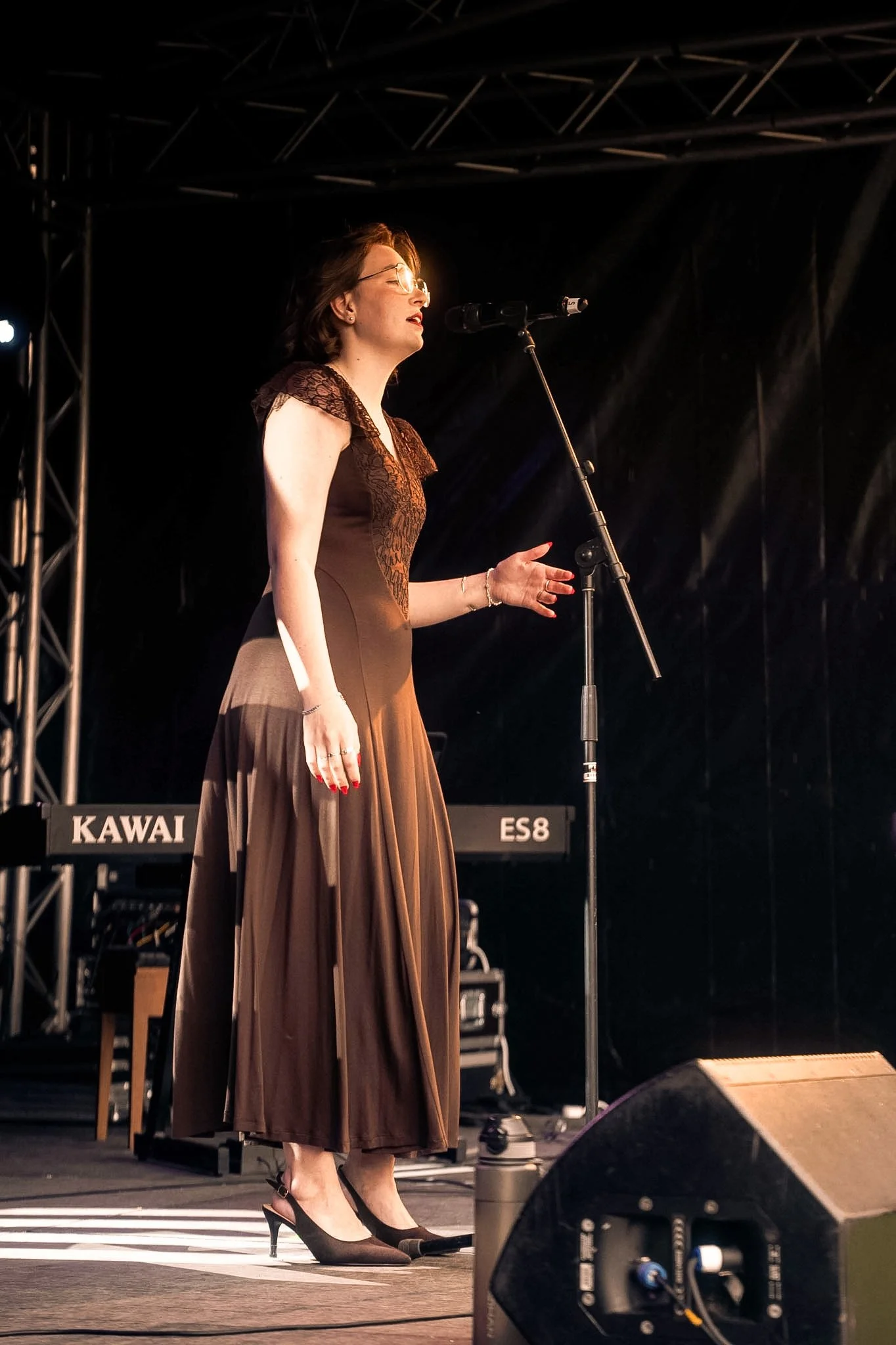 A woman singing into a microphone on stage, wearing glasses, a dress with lace details, and black high heels, with a keyboard in the background and stage equipment visible.