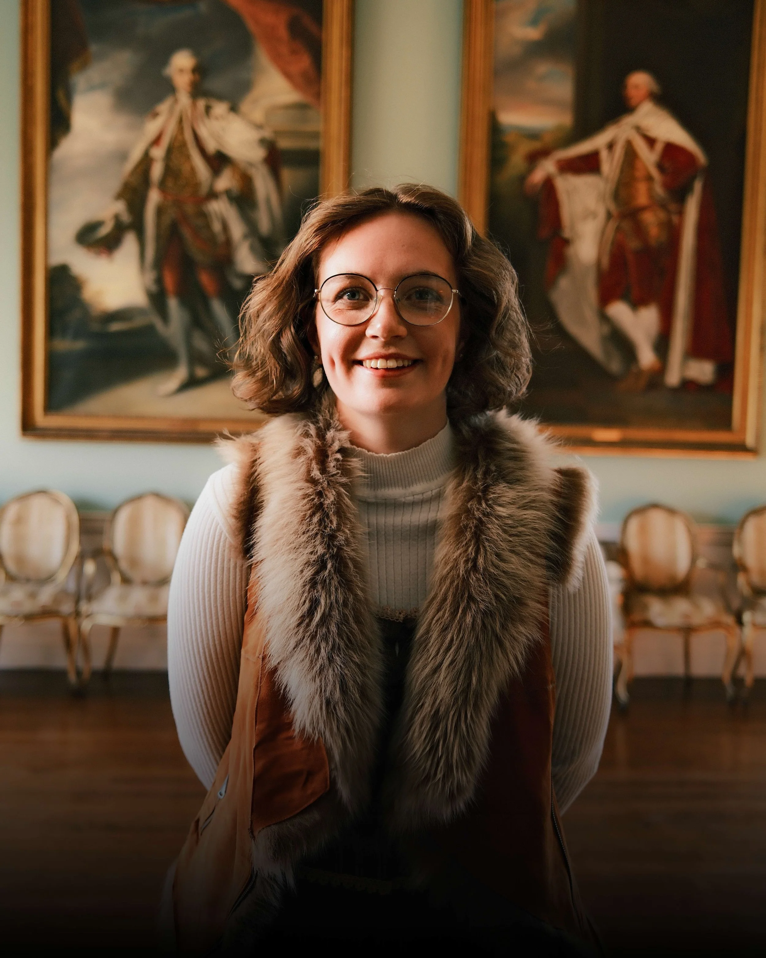 A smiling woman with glasses and curly hair stands in front of framed oil paintings of historical figures in an art gallery or museum. She wears a beige ribbed turtleneck sweater and a faux fur vest with a brown leather part.