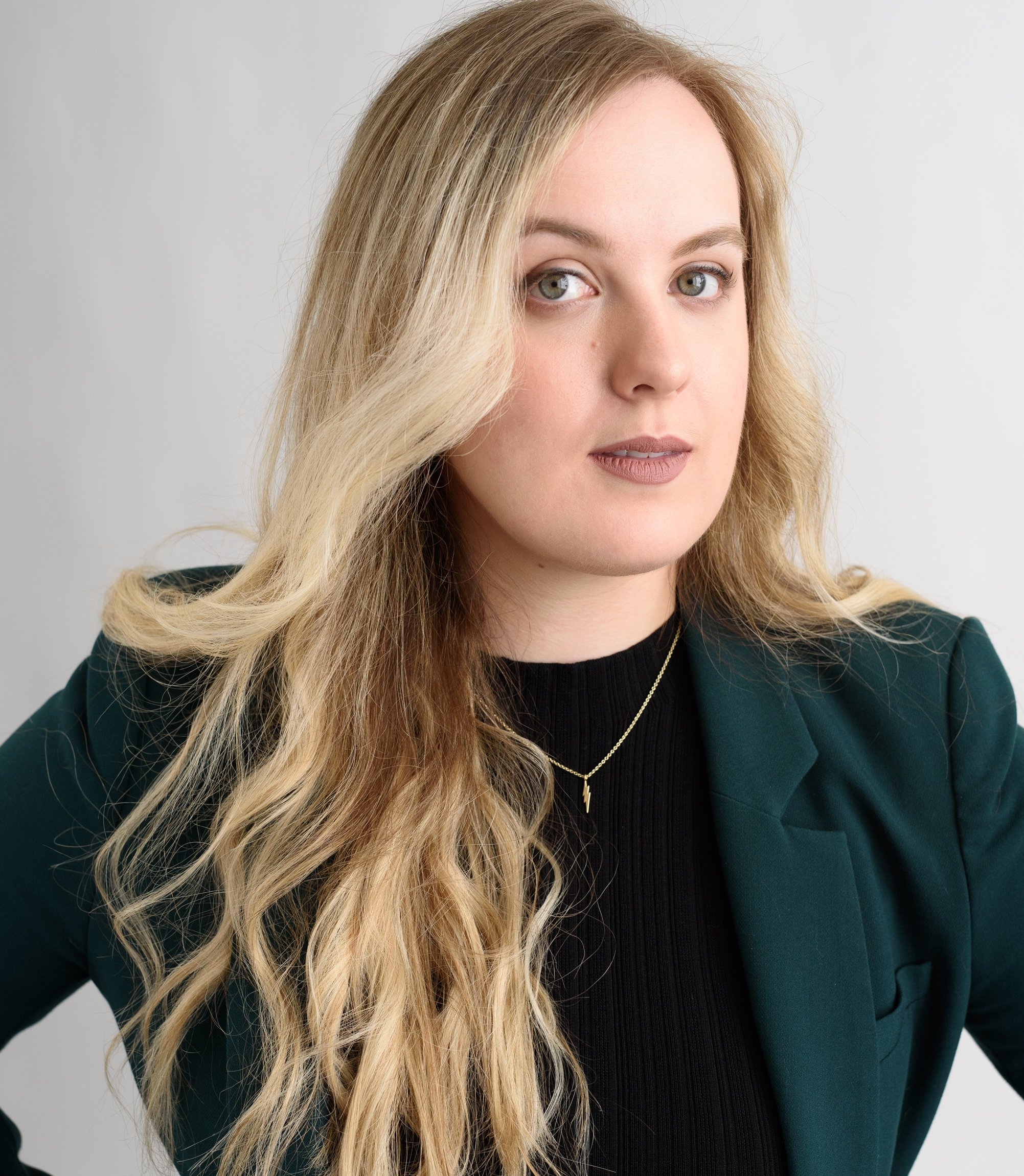 Close-up of a young woman with long wavy blonde hair, wearing a black top, gold necklace, and green blazer, against a plain white background.