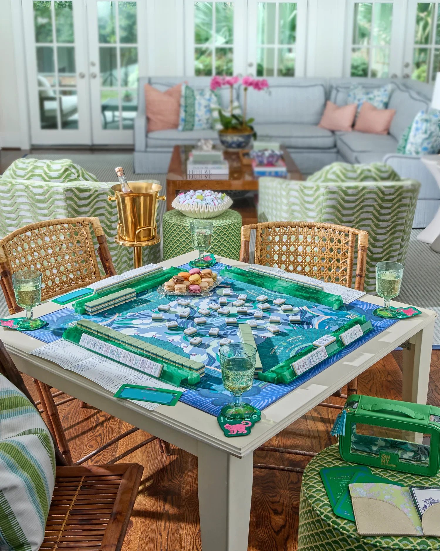 A lively indoor game room with a board game set up on a white table, surrounded by green and wicker chairs. The background features a sunlit sunroom with large windows, a cozy sofa, and a coffee table with books, a vase with pink flowers, and decorative pillows. The room has hardwood floors and a bright, inviting atmosphere.