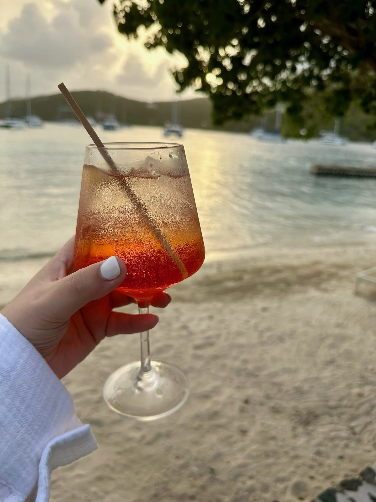 A glass of iced beverage with a straw, held by a person with white nail polish, in front of a beach scene with boats, water, trees, and a cloudy sky.