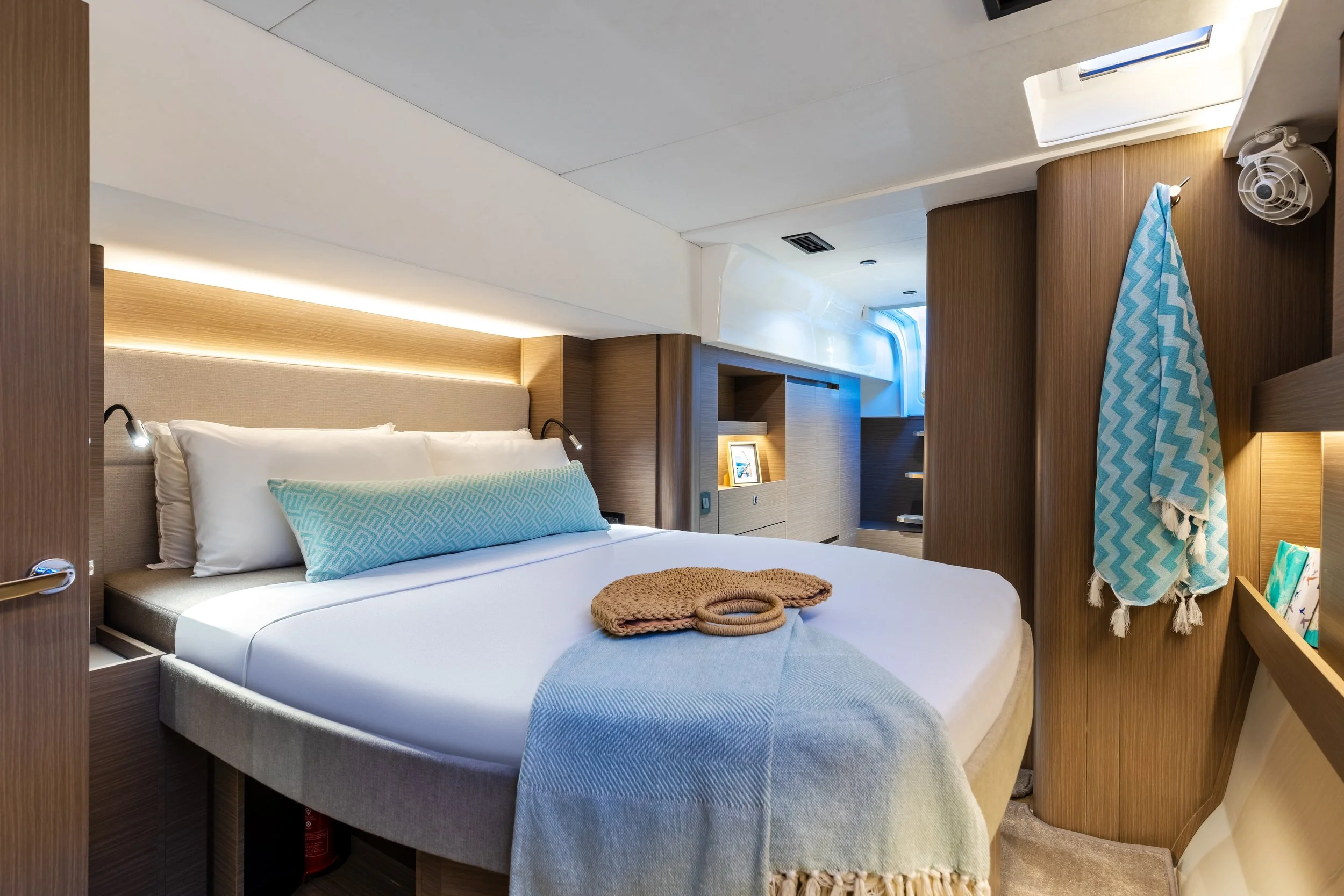 Interior of a modern yacht's cabin with a bed, wooden accents, reading lights, and a window with natural light.
