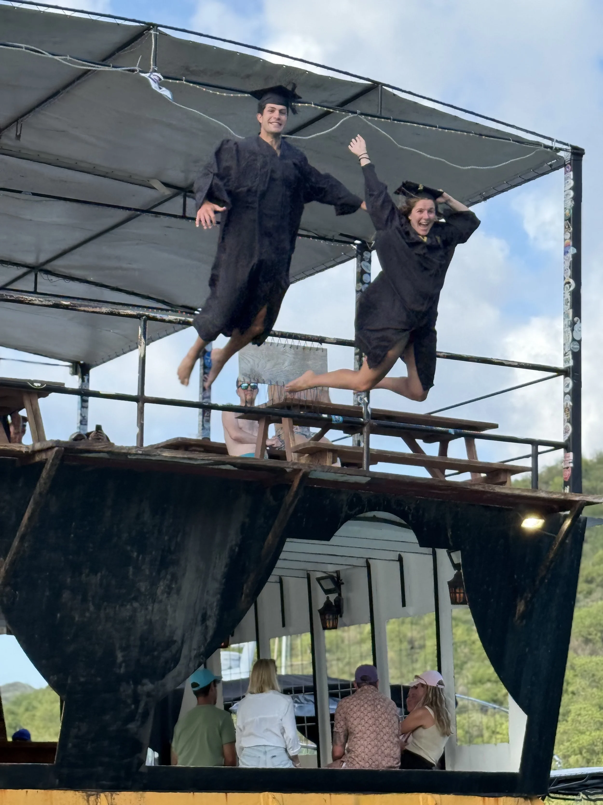 Graduates in caps and gowns jumping off of a floating platform into the ocean under a clear blue sky.