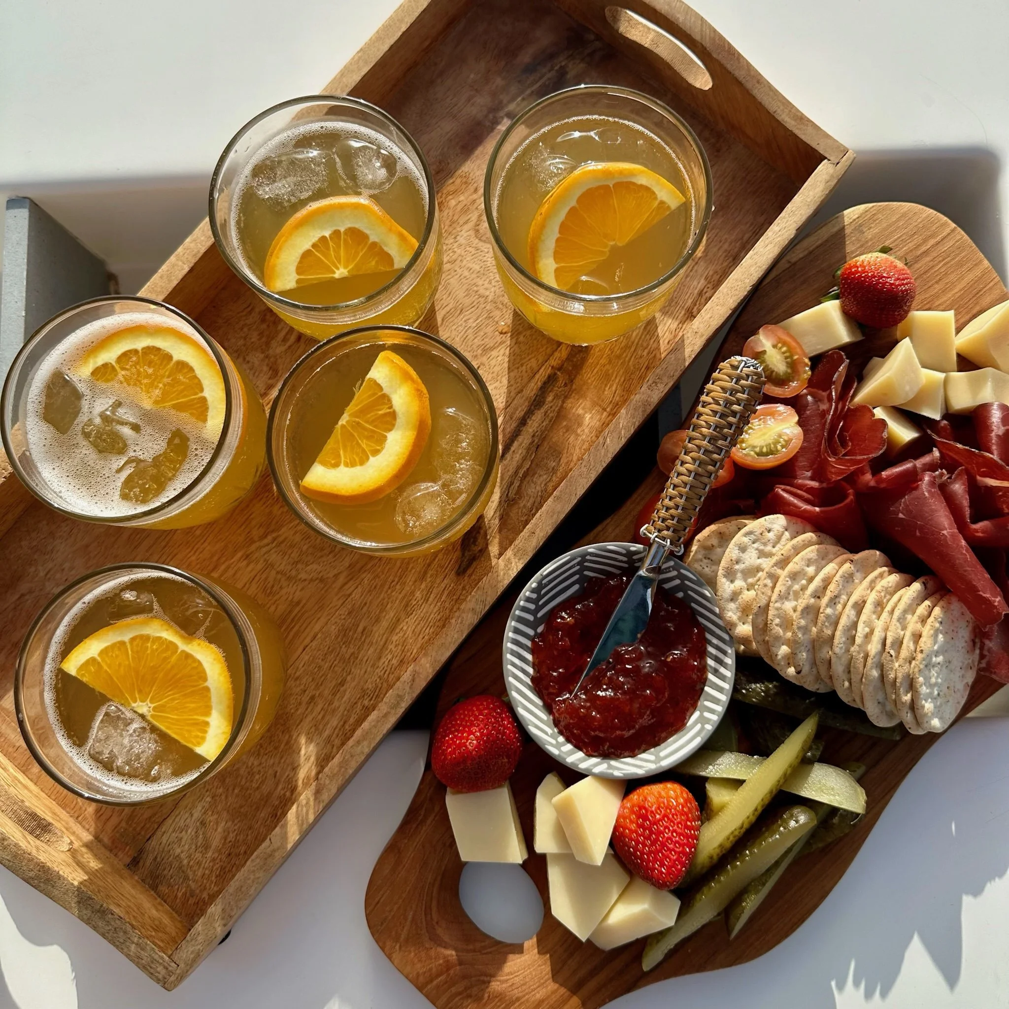 Overhead view of a wooden serving tray with glasses of sparkling juice garnished with orange slices, alongside an assortment of cheeses, strawberries, grapes, tomato slices, and crackers on a white surface.