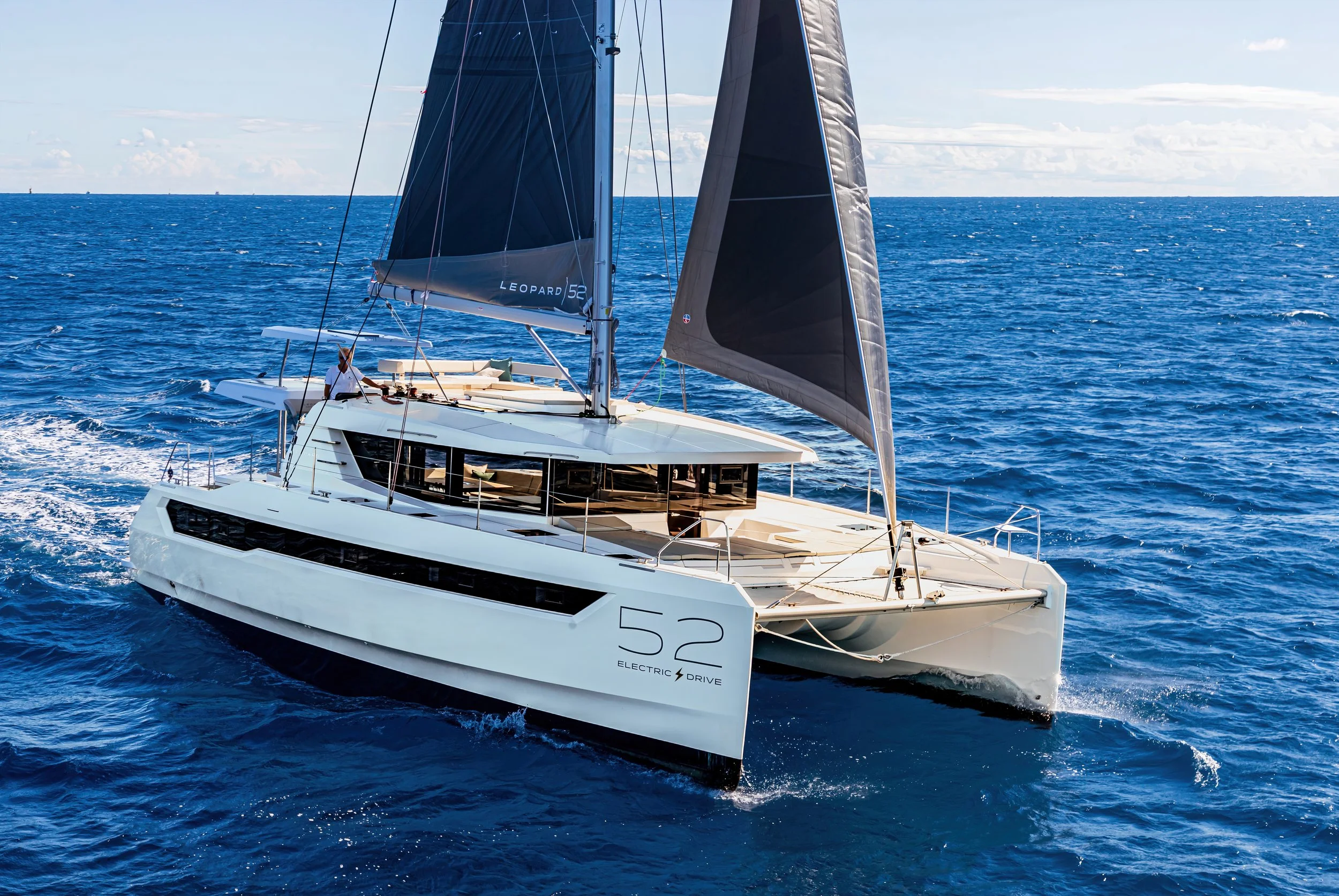 A modern sailing yacht on the open ocean with black, blue, and gray sails, and a crew member on deck, under a partly cloudy sky.
