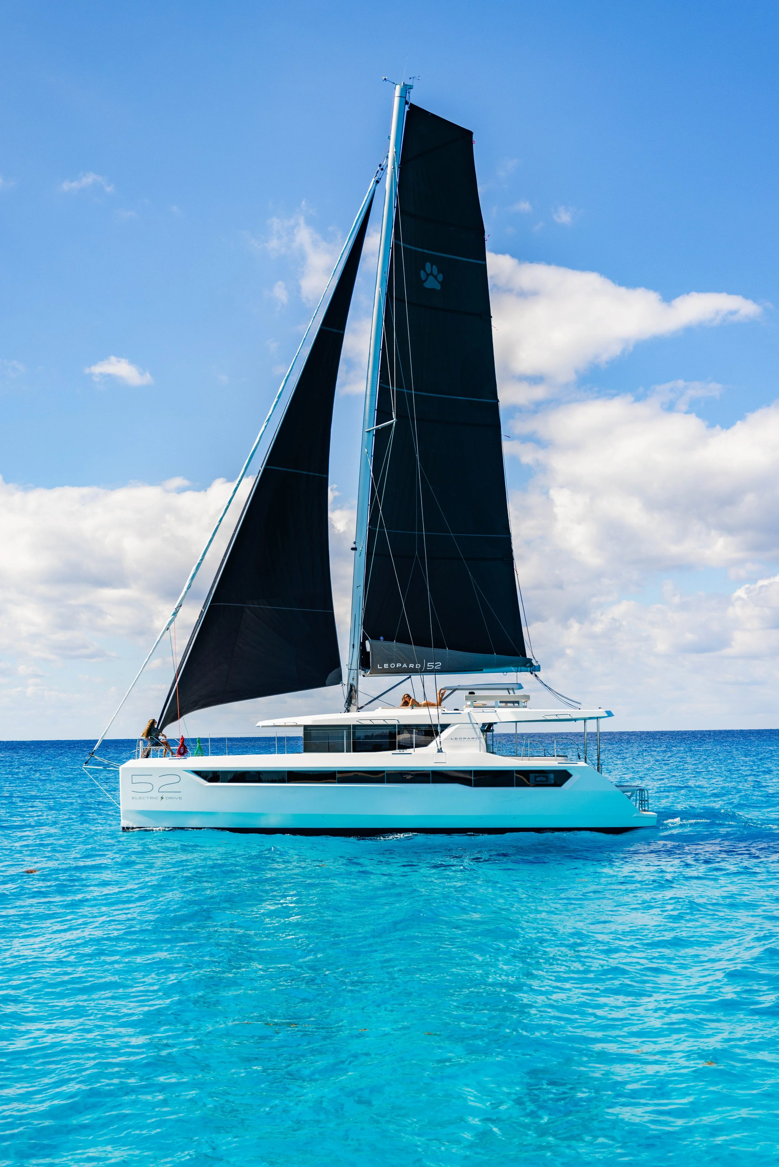 A modern white catamaran yacht with navy sails, sailing on clear blue ocean water under a partly cloudy sky.