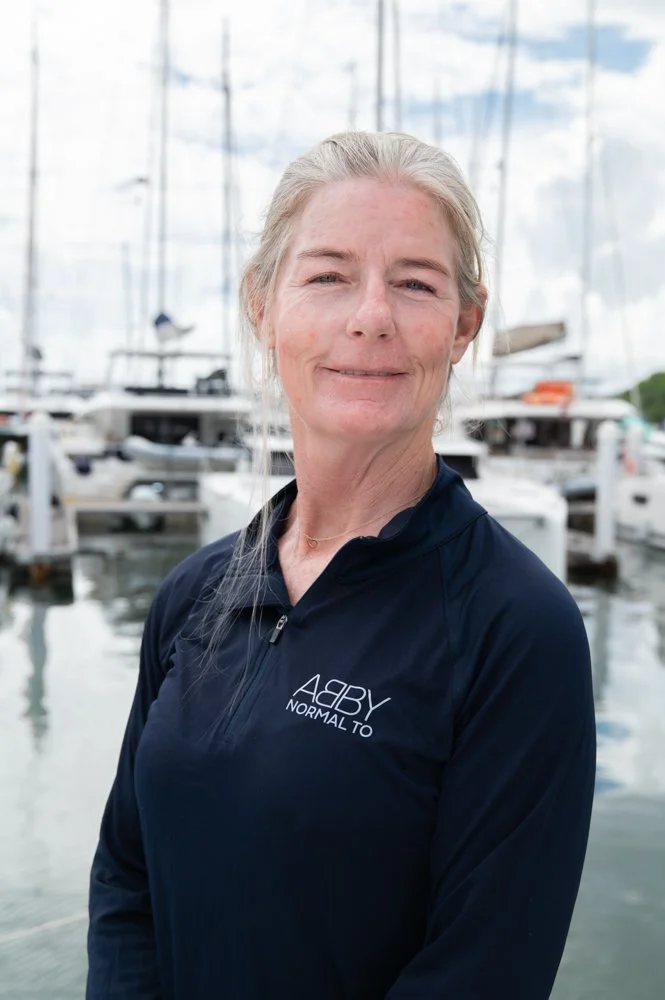 A woman with light gray hair in a ponytail, smiling, standing near boats on a marina, wearing a dark navy athletic jacket that says "ABBY NORMAL TO".